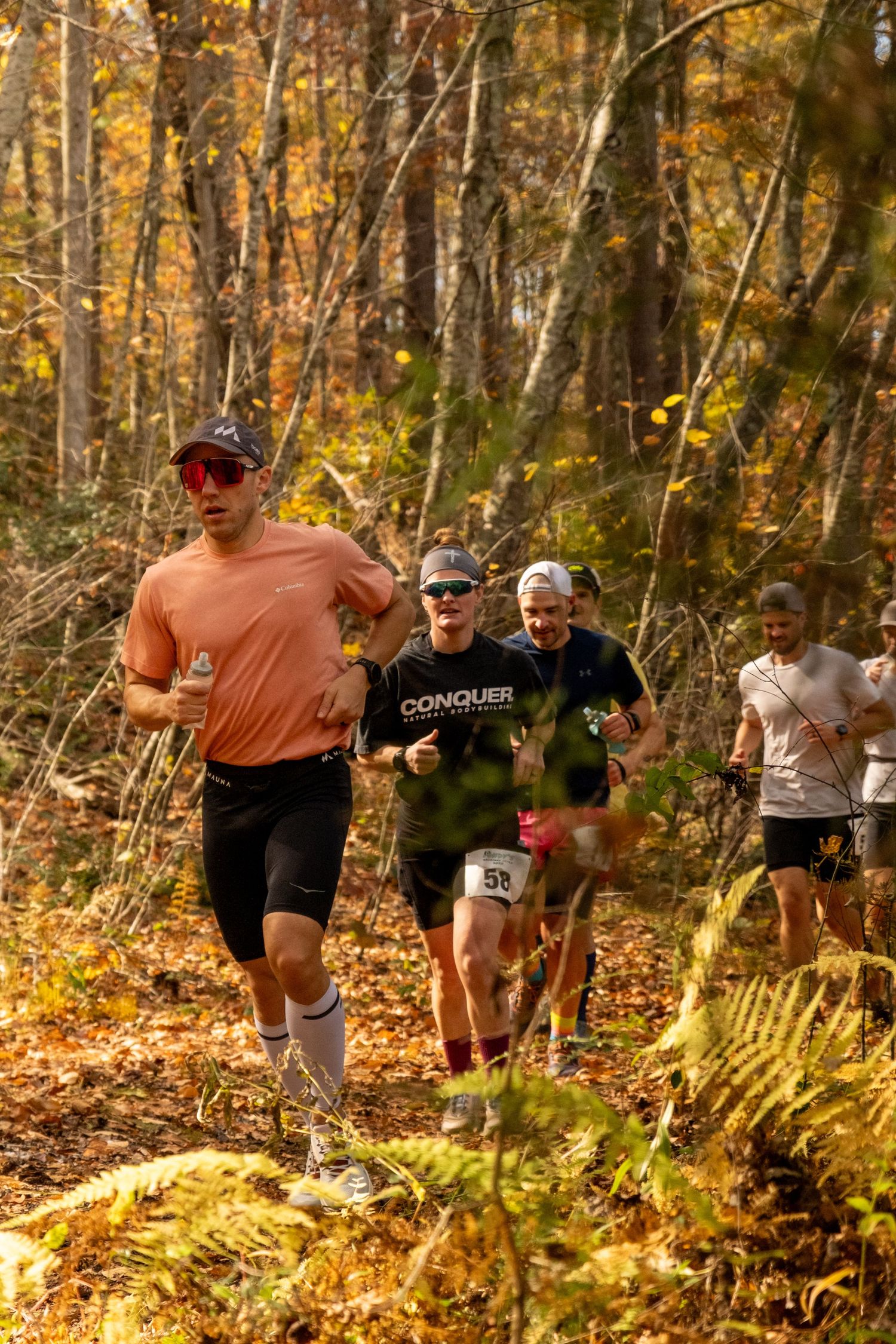 Trail runners navigate through a scenic autumn forest with golden foliage and ferns during a cross-country race.