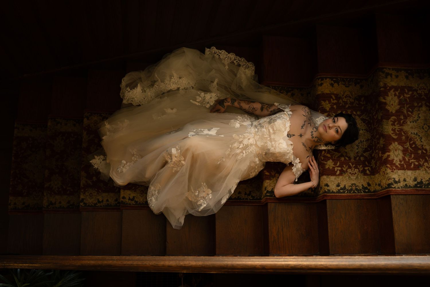 Woman in flowing champagne lace gown dramatically posed on wooden stairs in ethereal bridal fashion photography.