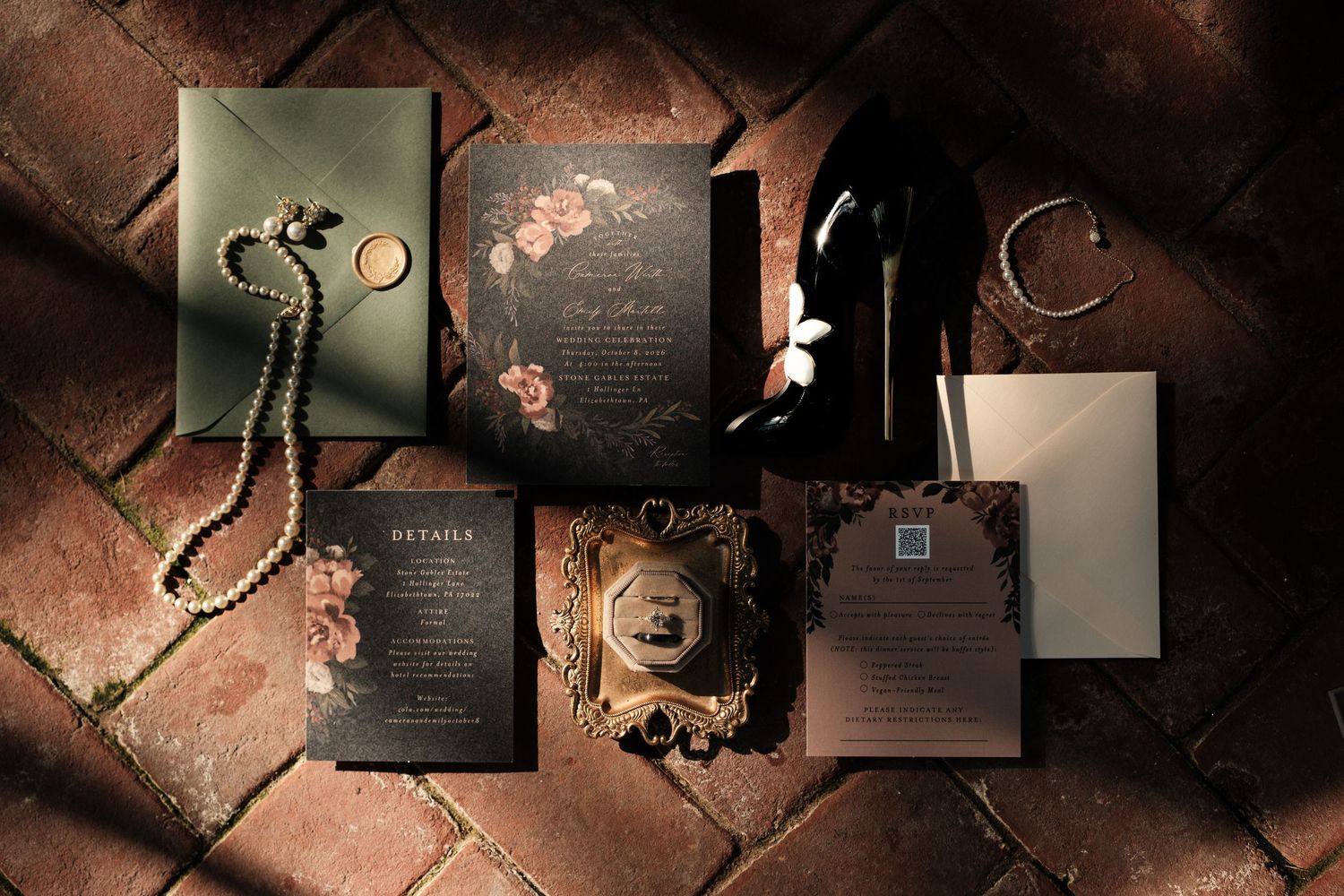 Moody floral wedding invitation suite with pearl necklace, wax seal, gold frame and black heels on rustic brick flooring.