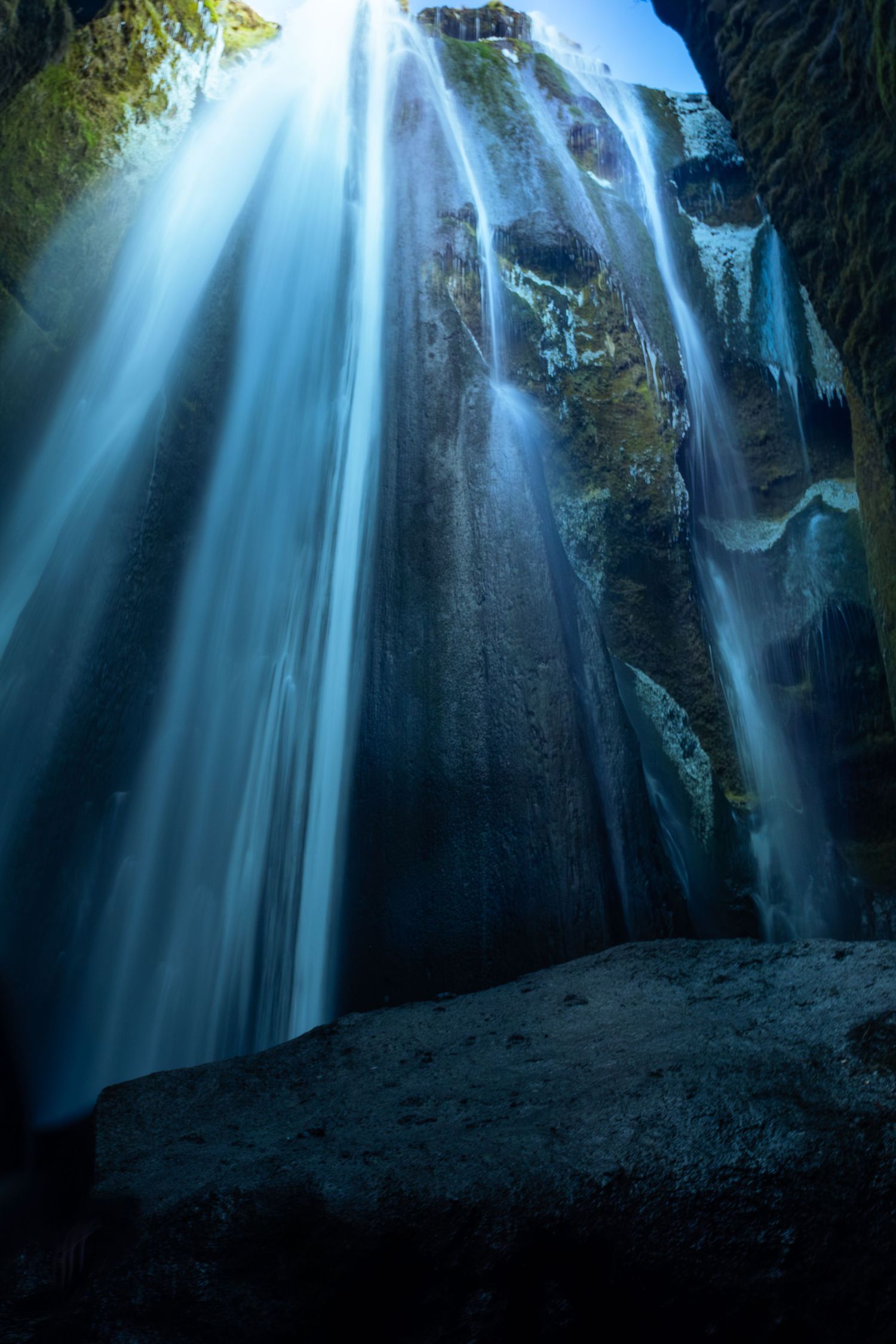 Dramatic waterfall cascades down dark cave walls with ethereal blue light filtering through the cavern opening.