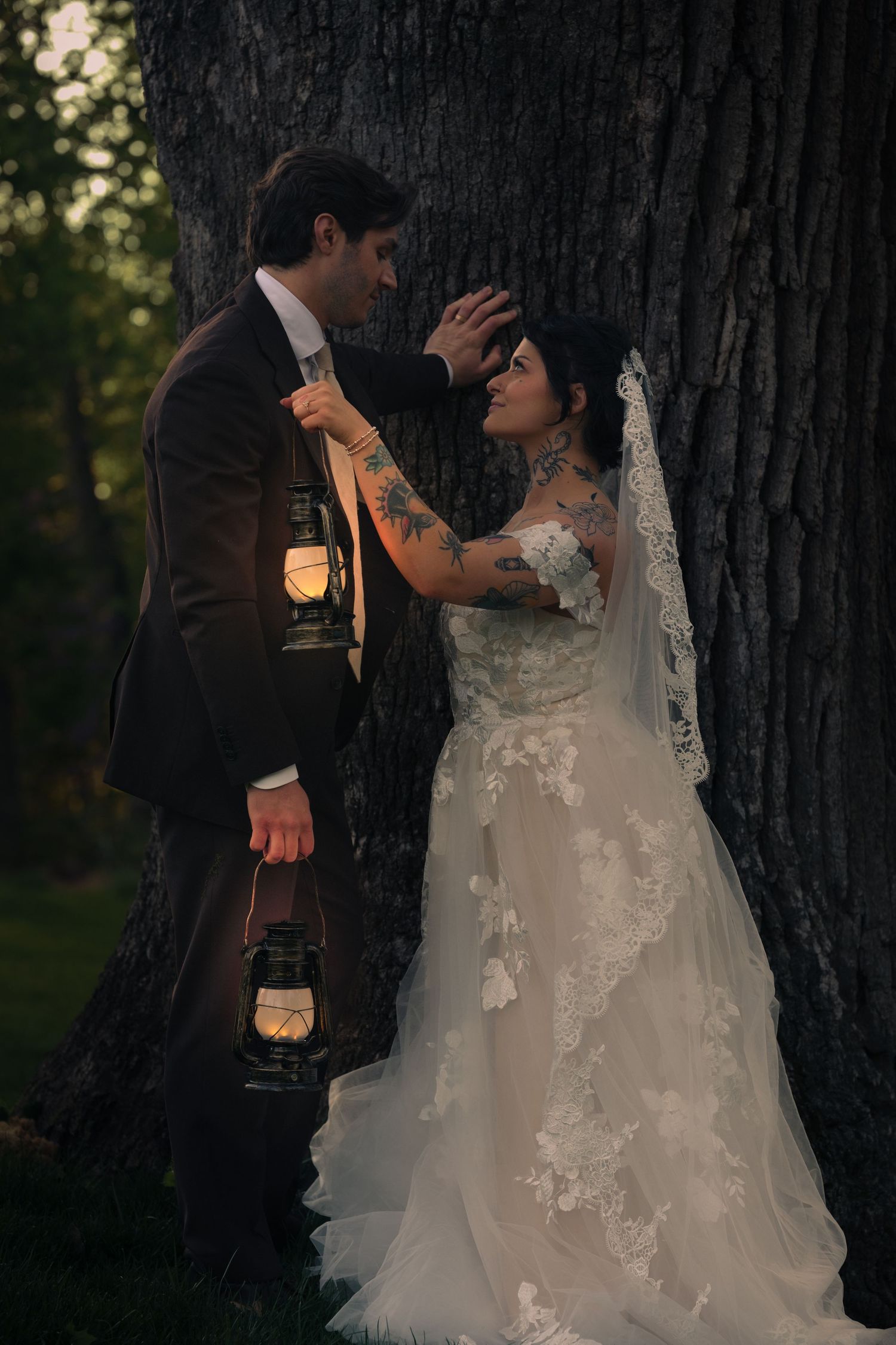 Bride and groom lean against a large tree at dusk, groom holding a vintage lantern, bride in lace gown with veil and tattoos.