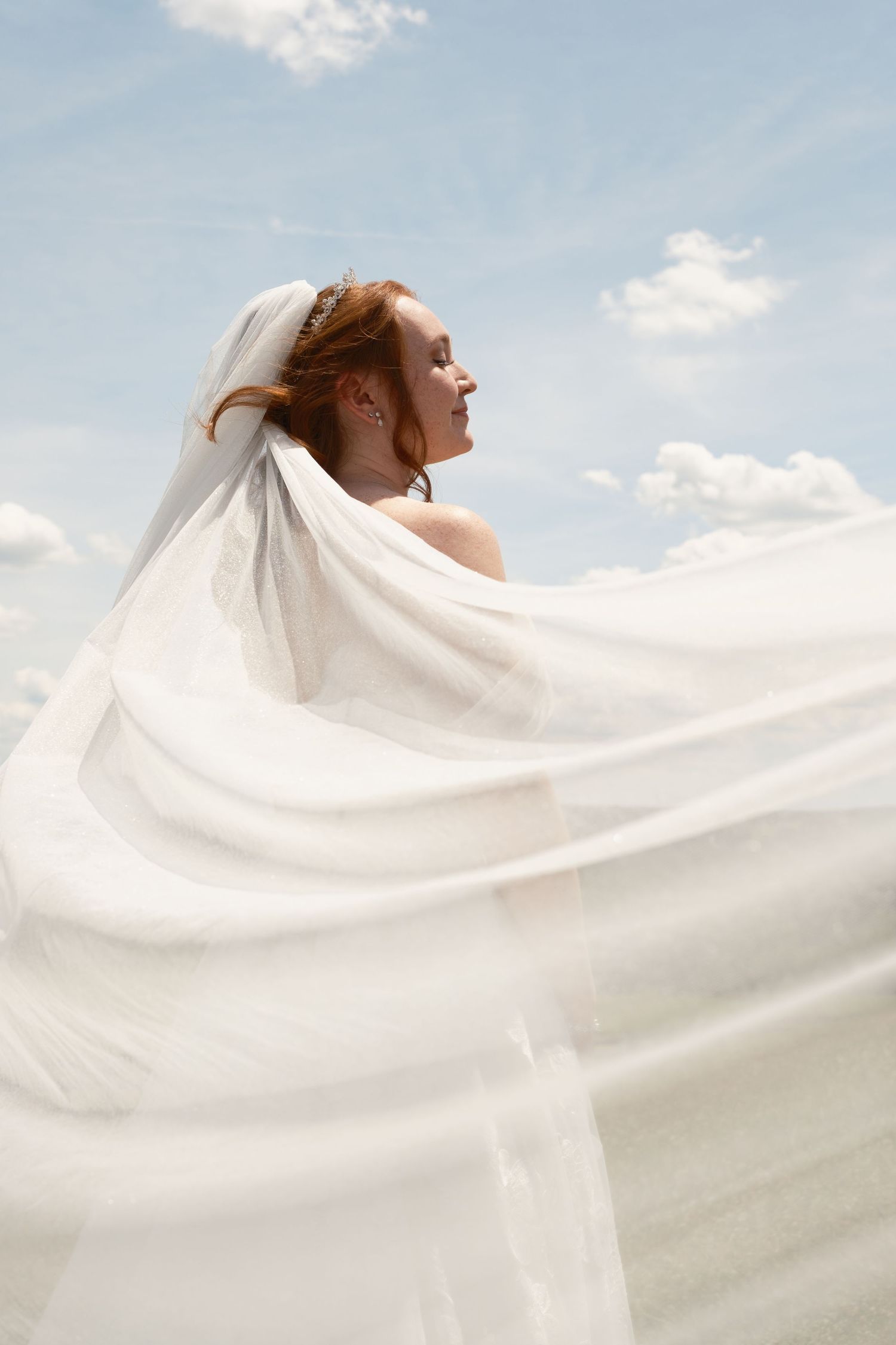 A bride in a flowing white veil stands outdoors against a blue sky, her red hair catching the breeze.