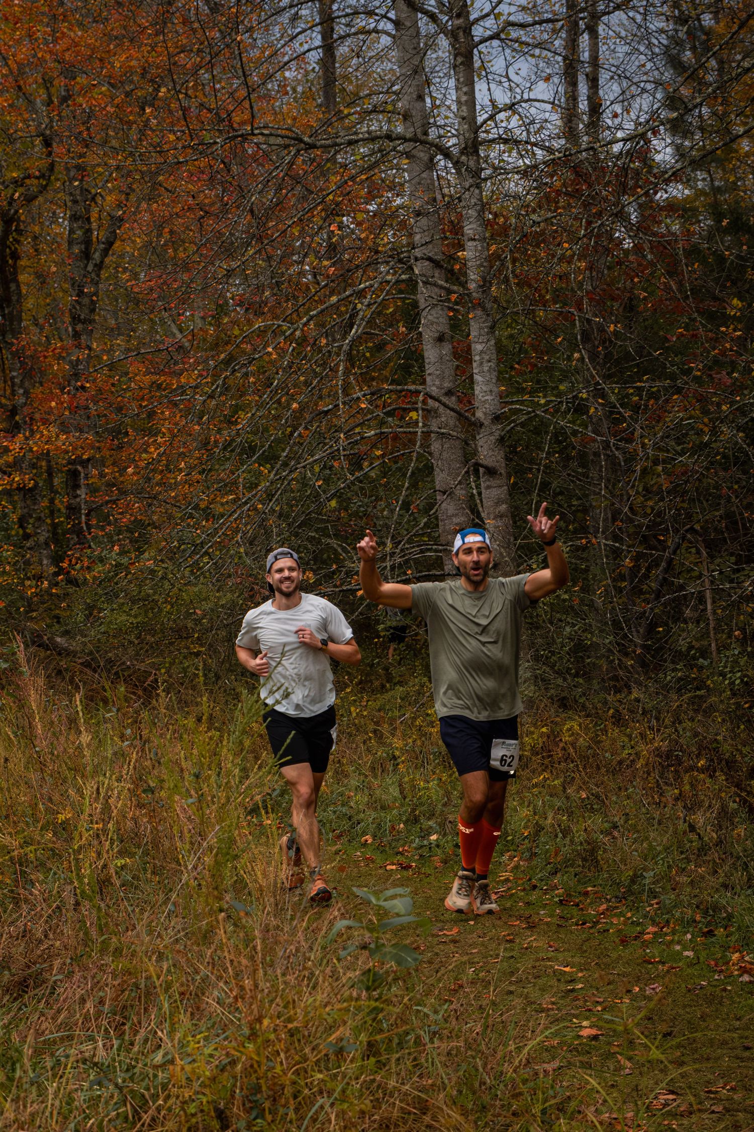 Two people jumping and posing playfully in autumn woods with orange and brown fall foliage in the background.