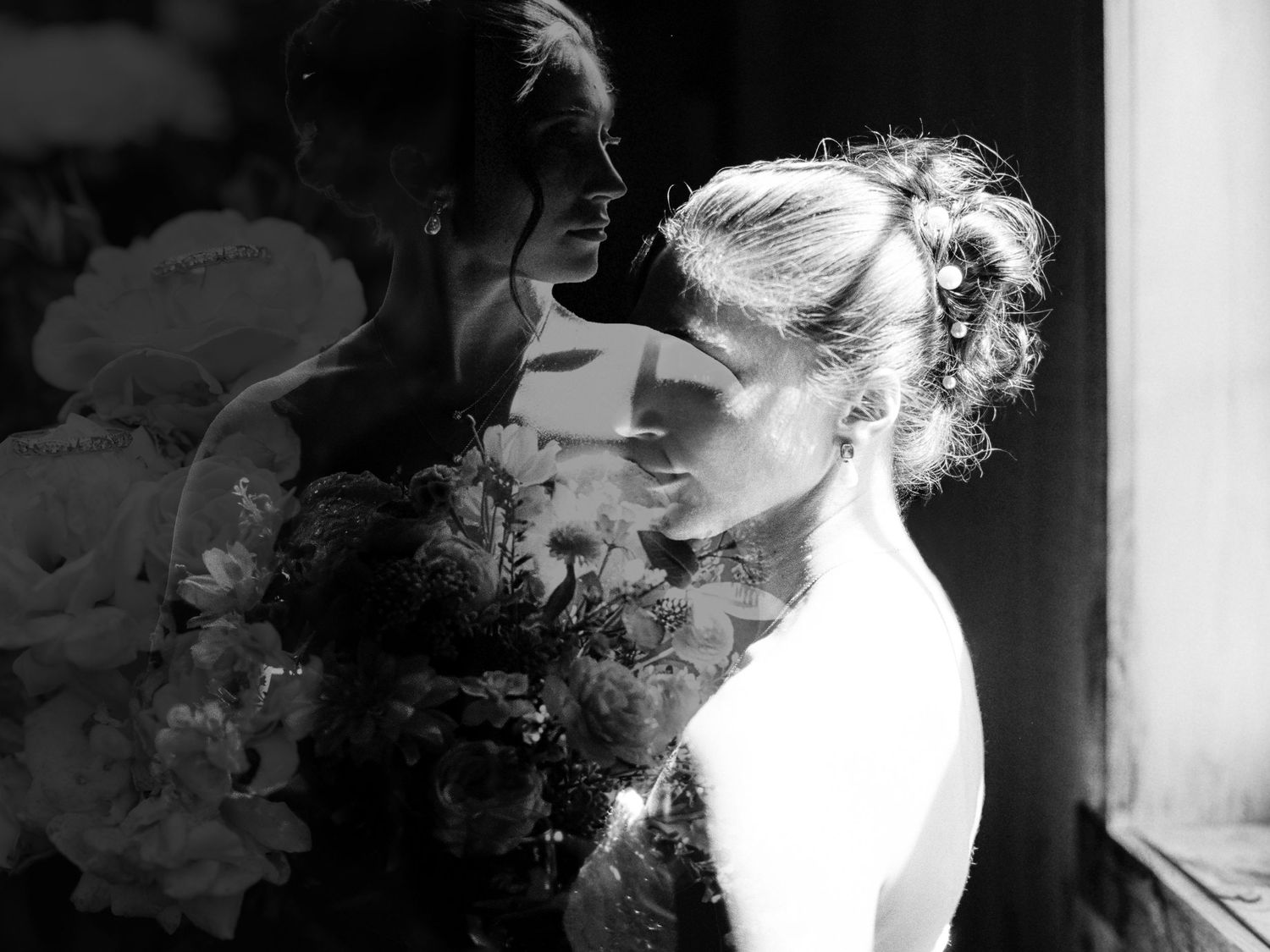 Black and white photo of two brides in wedding gowns sharing an intimate moment with a floral bouquet.