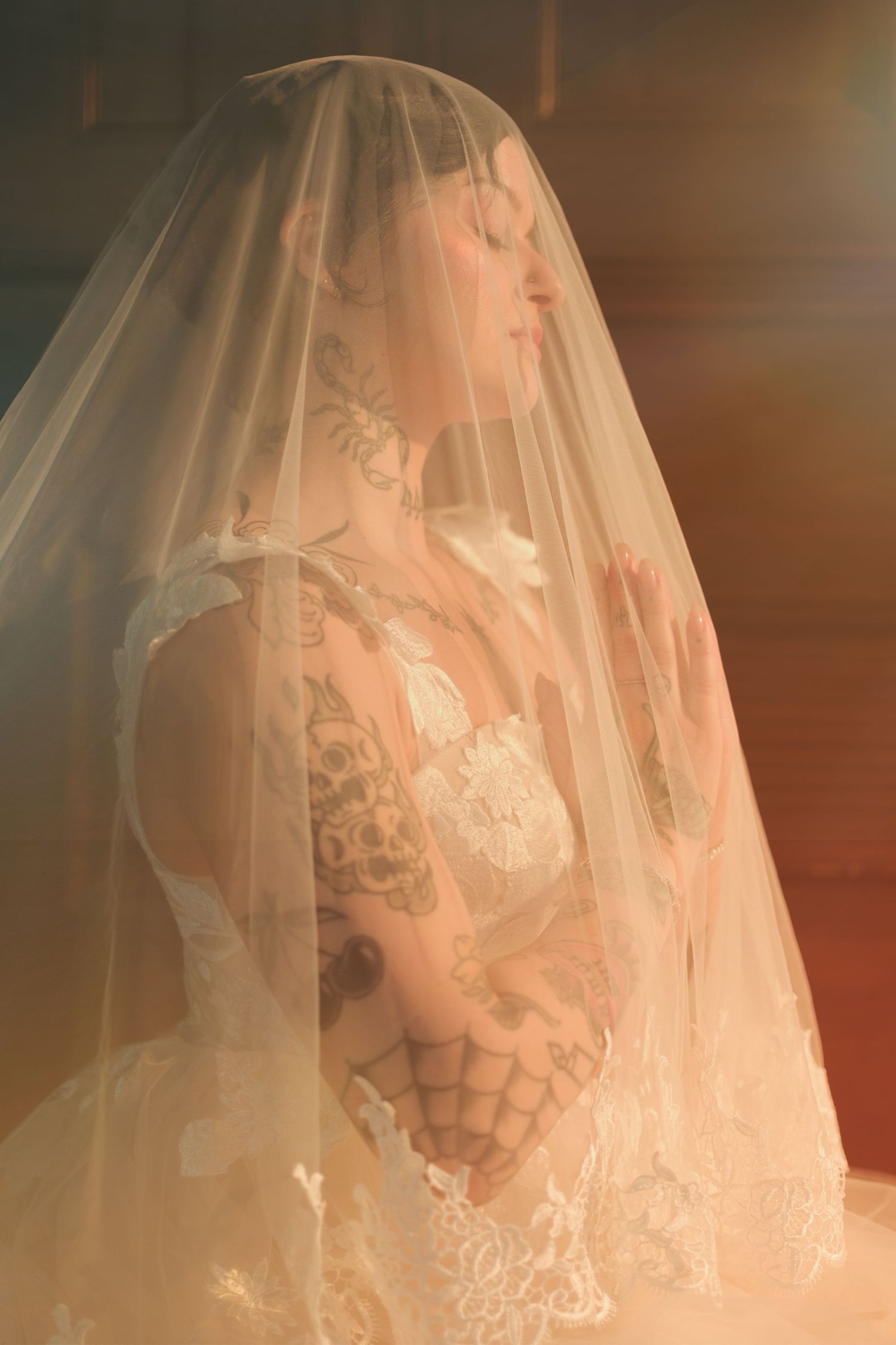 Tattooed bride in lace wedding dress with veil draped over head, hands clasped in prayer pose, warm golden light.