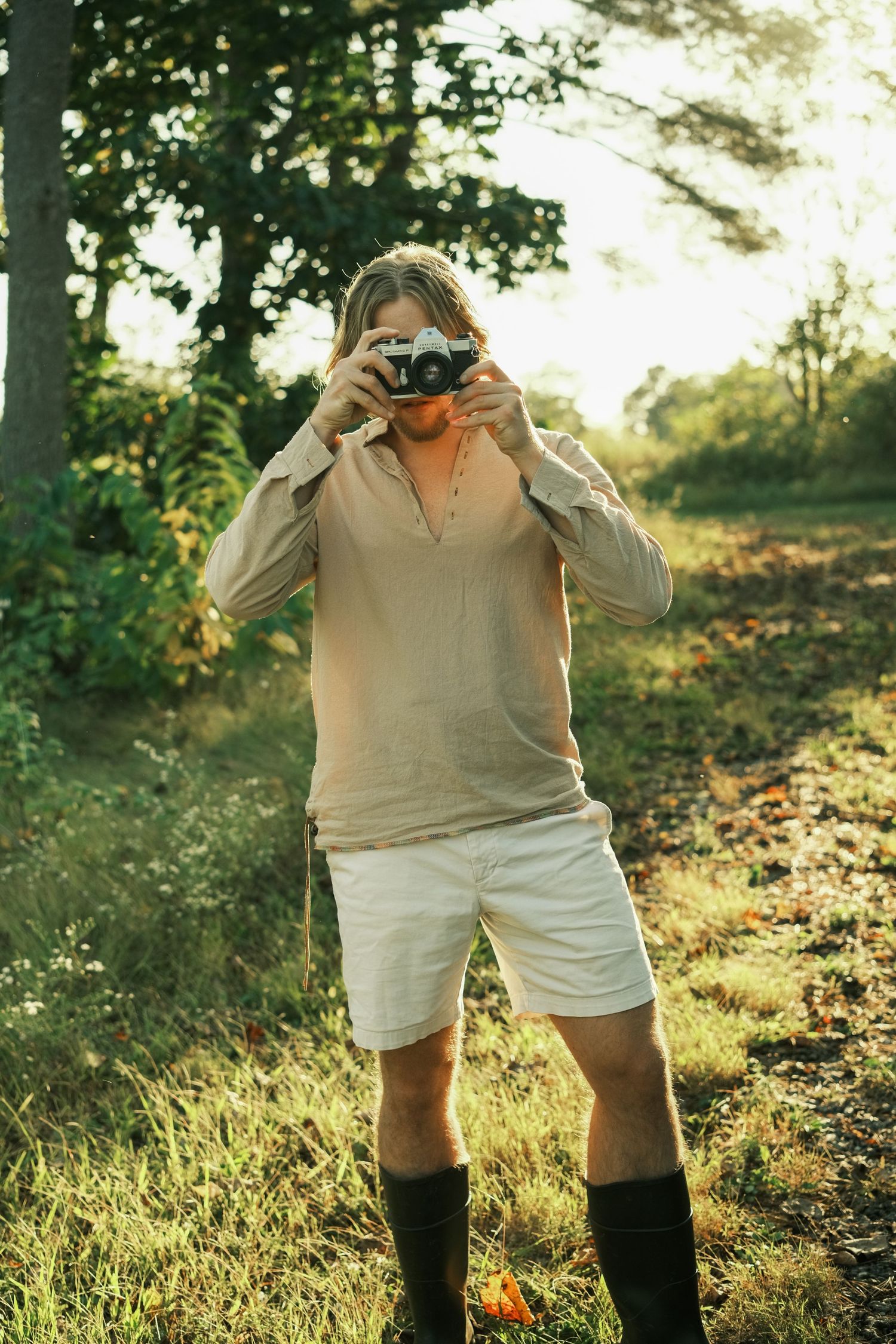 Person with long hair photographing outdoors in beige shirt, white shorts, and black boots in a sunlit forest clearing.