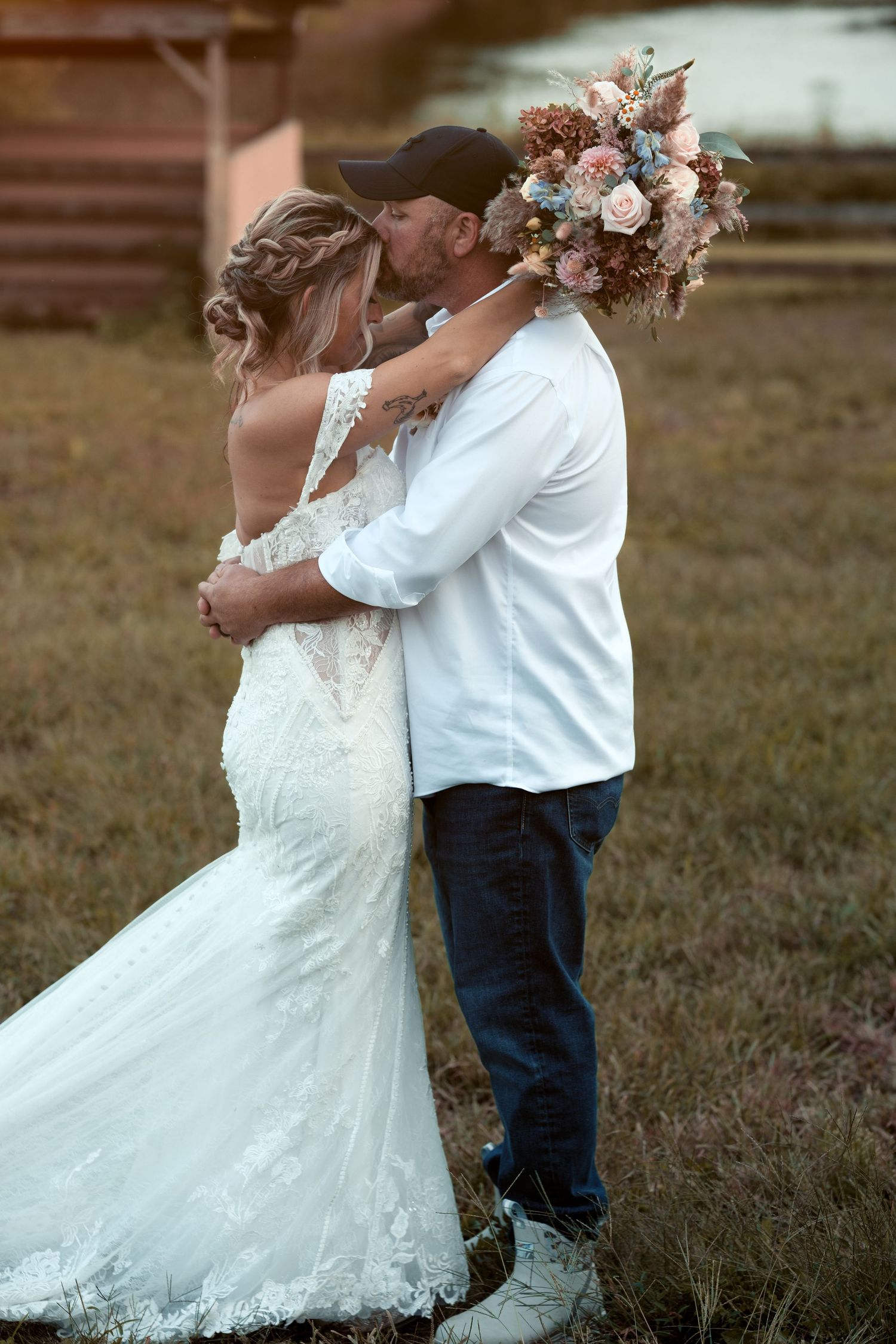 Bride in a lace wedding gown embraces and kisses a groom in a cap while holding a pastel bouquet in a grassy field at sunset.