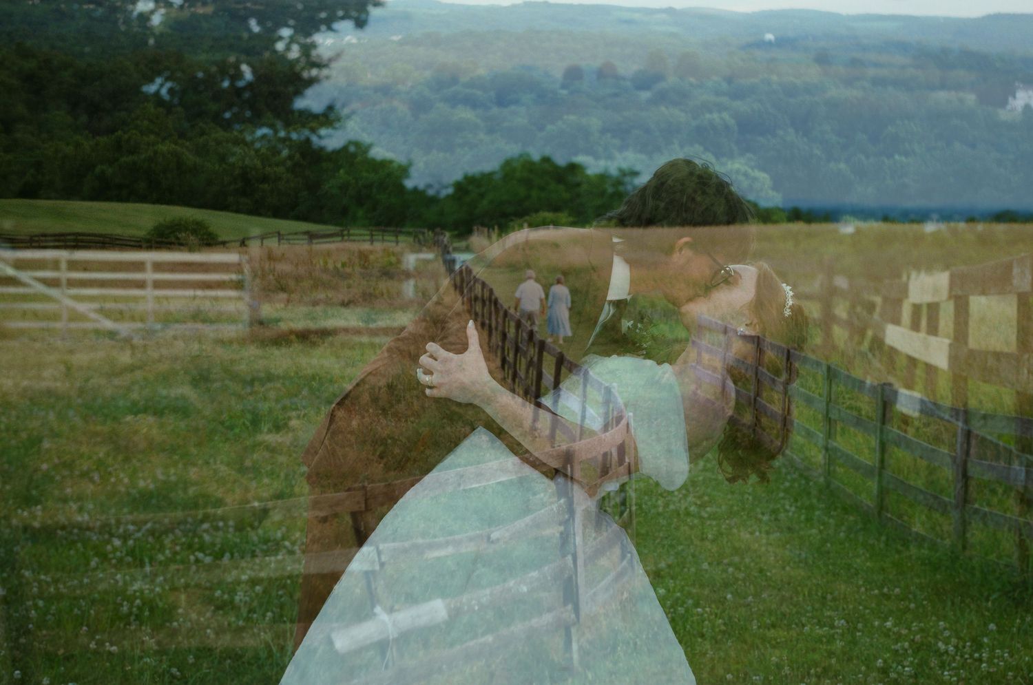 Double exposure photo blending a couple embracing with a scenic rural countryside landscape featuring green fields and mountains.