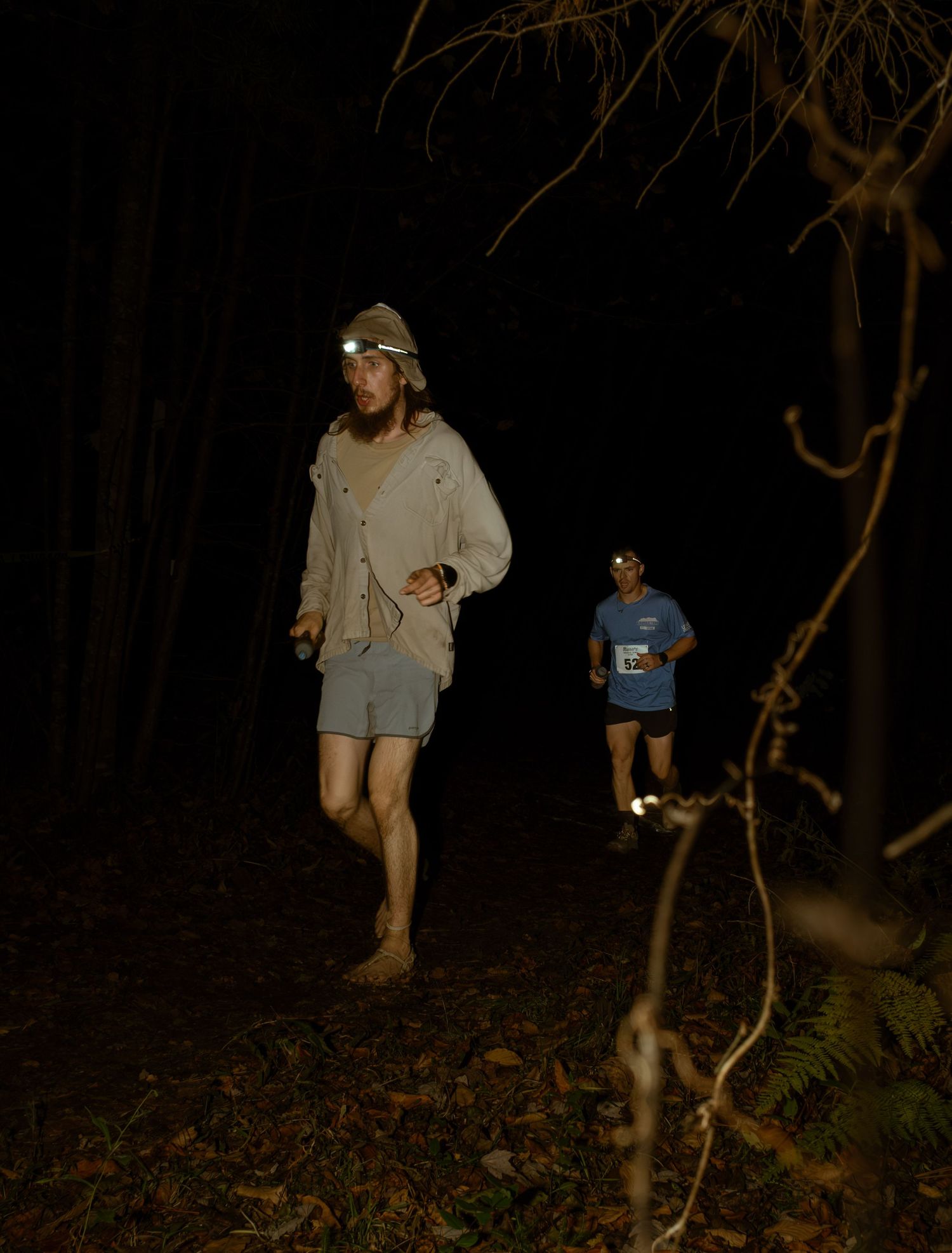 People explore a dark wooded area at night with flashlights casting eerie shadows among bare tree branches.