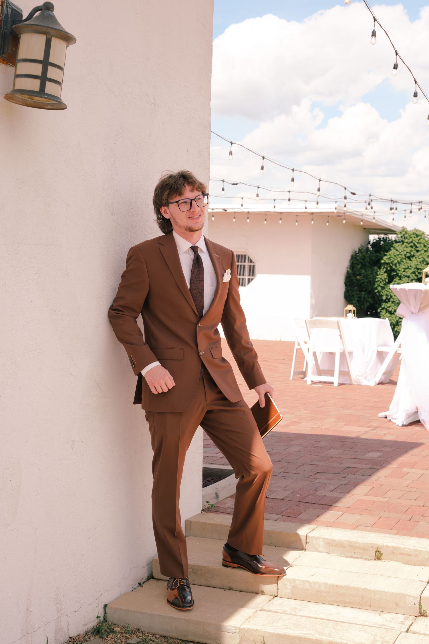 Groom in brown suit leaning against white wall at outdoor wedding venue with string lights.