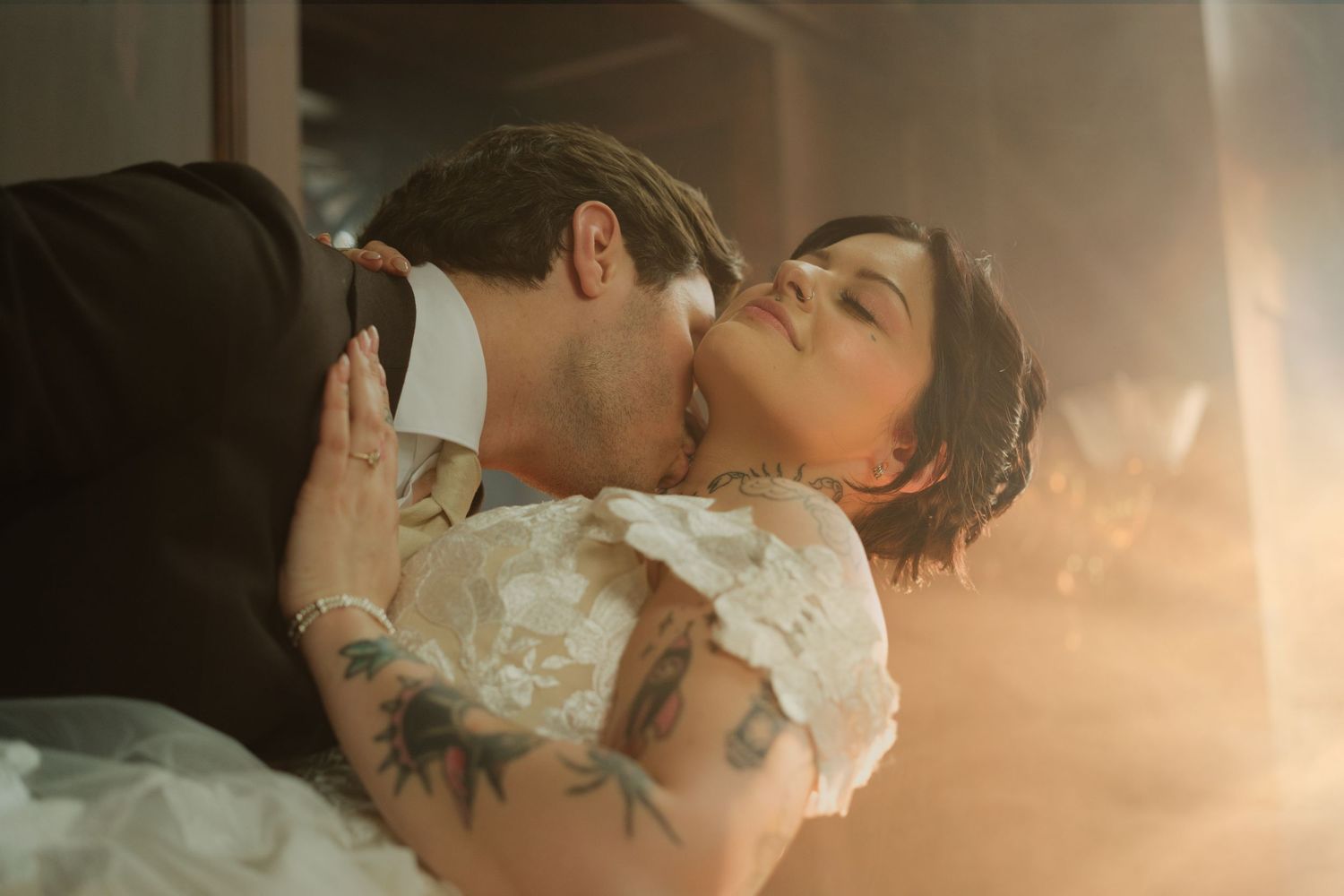 A couple shares a romantic kiss, bride in lace dress with tattoos, groom in suit, soft golden light.