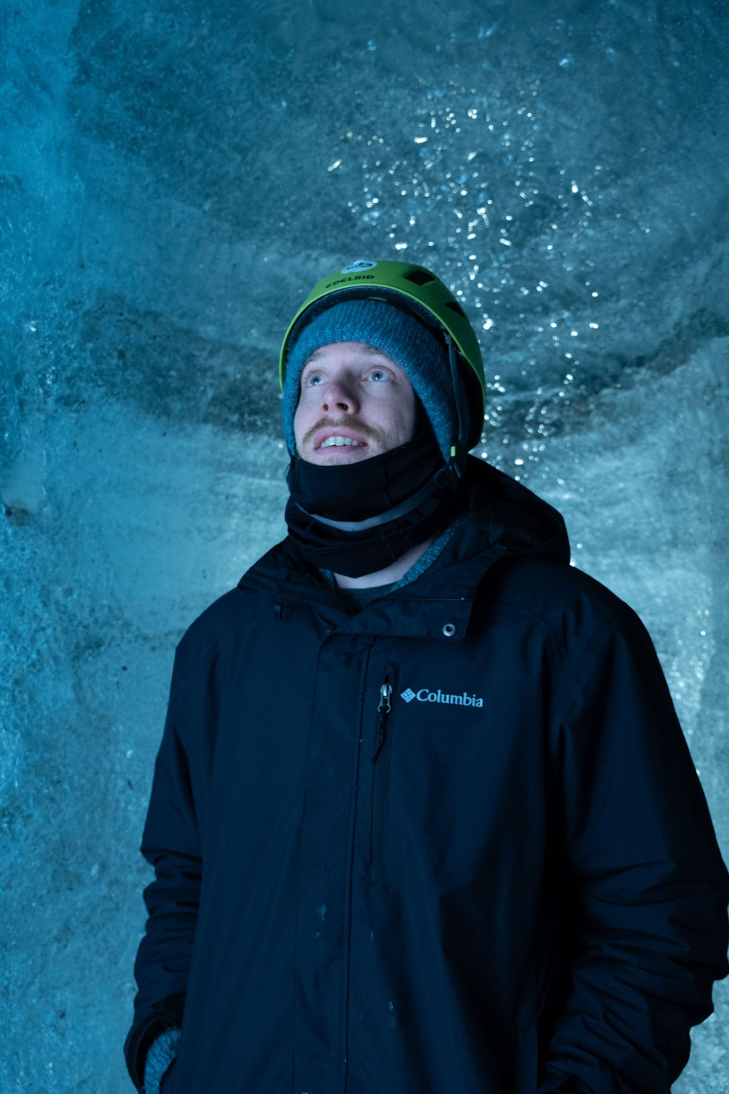 Person wearing black Columbia jacket and winter hat explores inside an icy blue glacial cave.