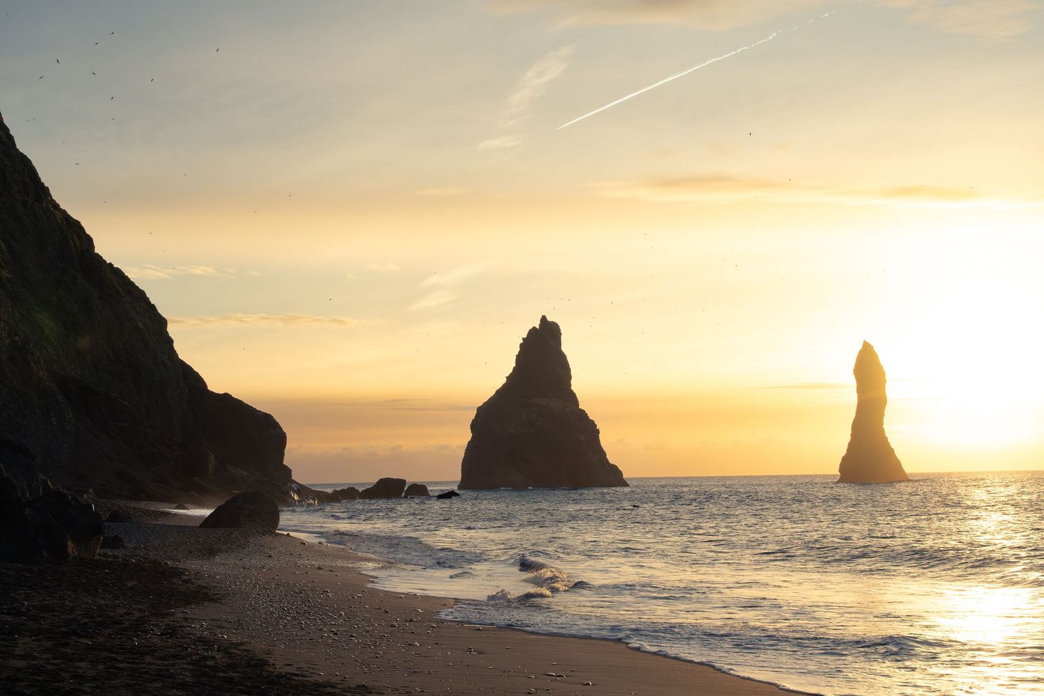 Dramatic sunset illuminates rocky sea stacks and coastal cliffs along a rugged beach shoreline.
