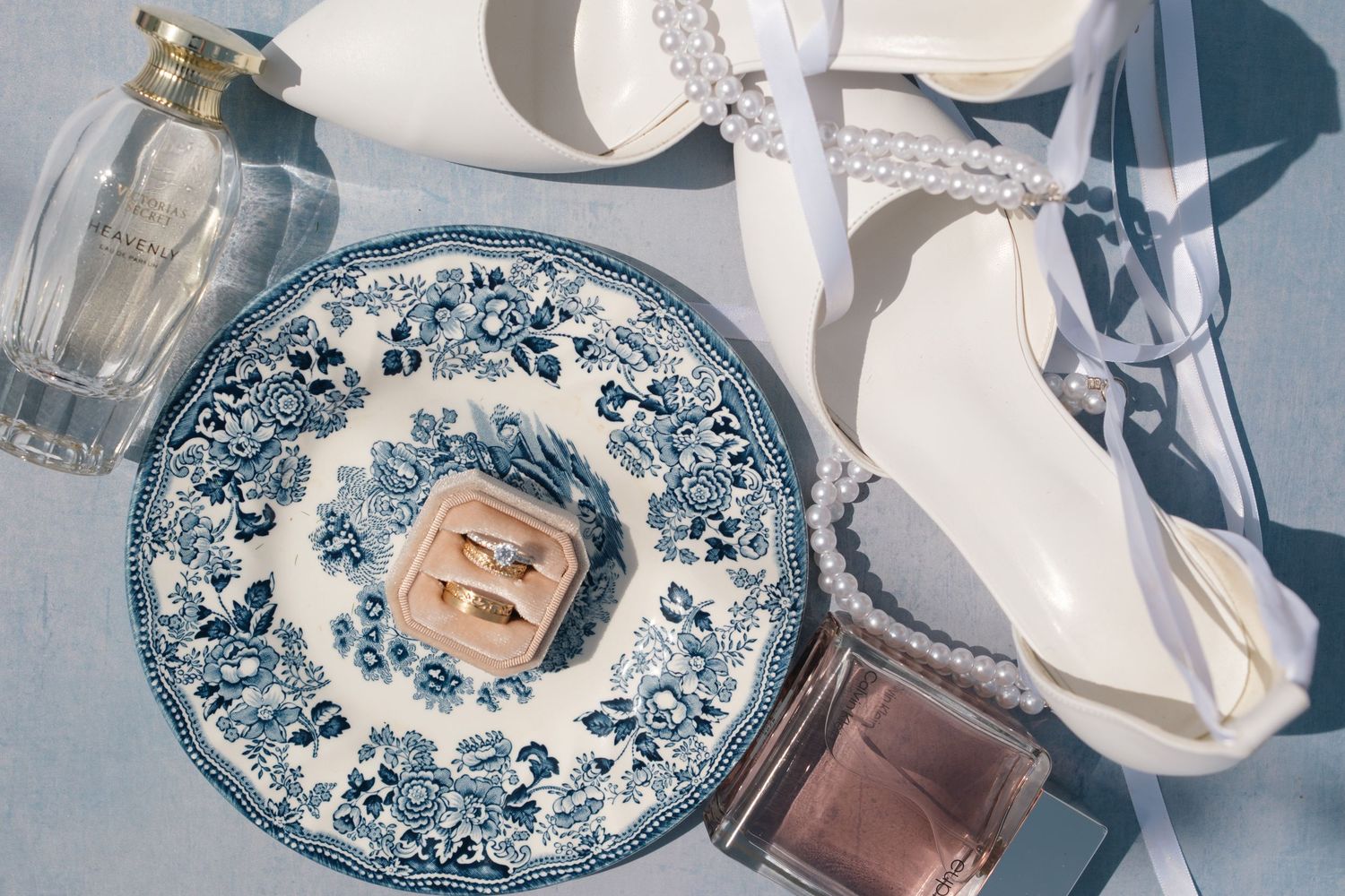 Blue and white floral plate with gold wedding rings, white heeled bridal shoes with pearls, and perfume bottles on gray surface.