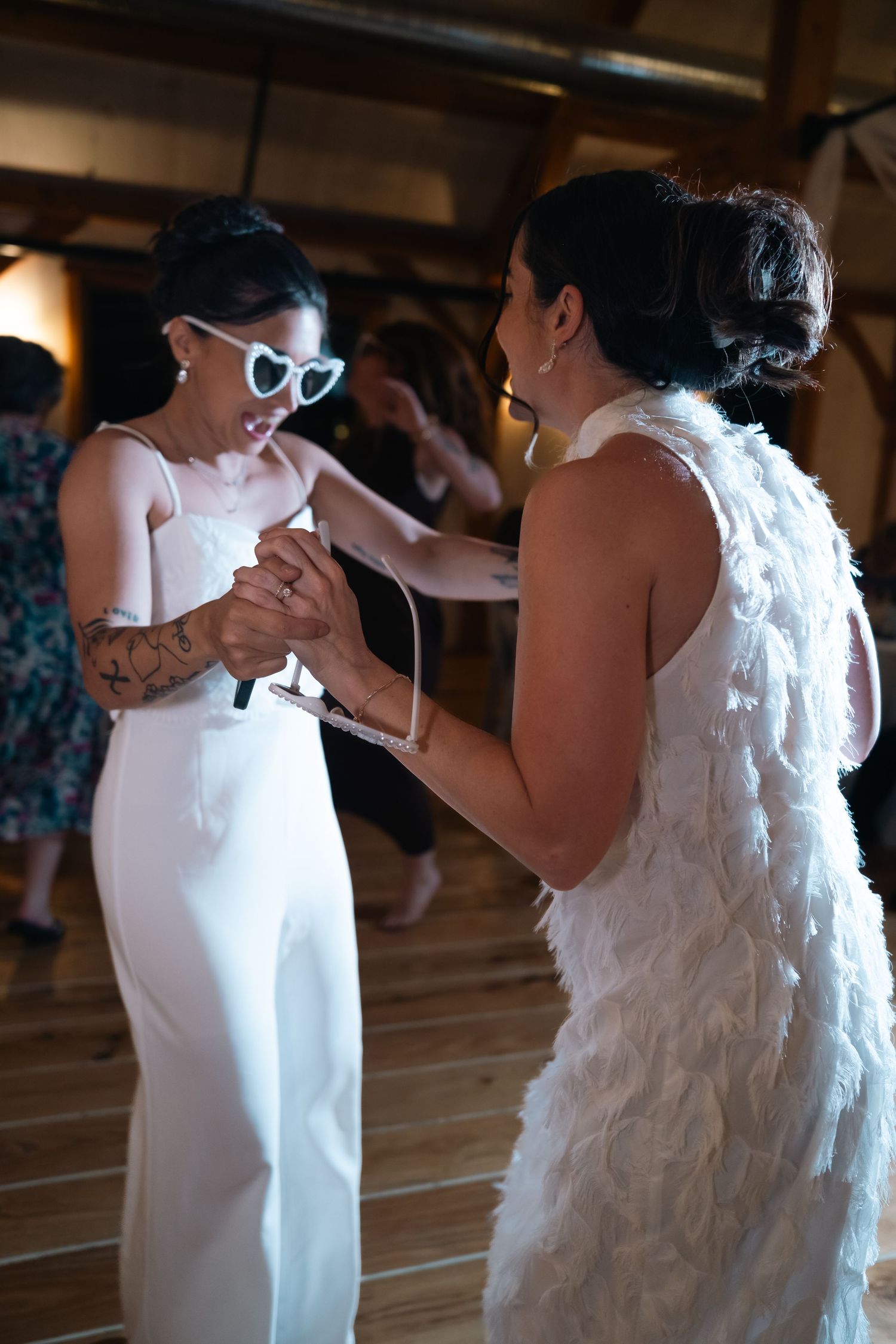 Two women in white dresses dancing together at wedding, one wearing heart-shaped sunglasses in barn venue.