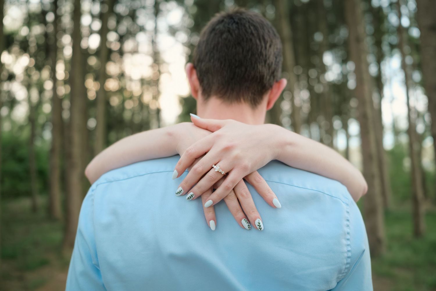 Couple embracing outdoors in a forest, woman's hand with engagement ring resting on man's shoulder.