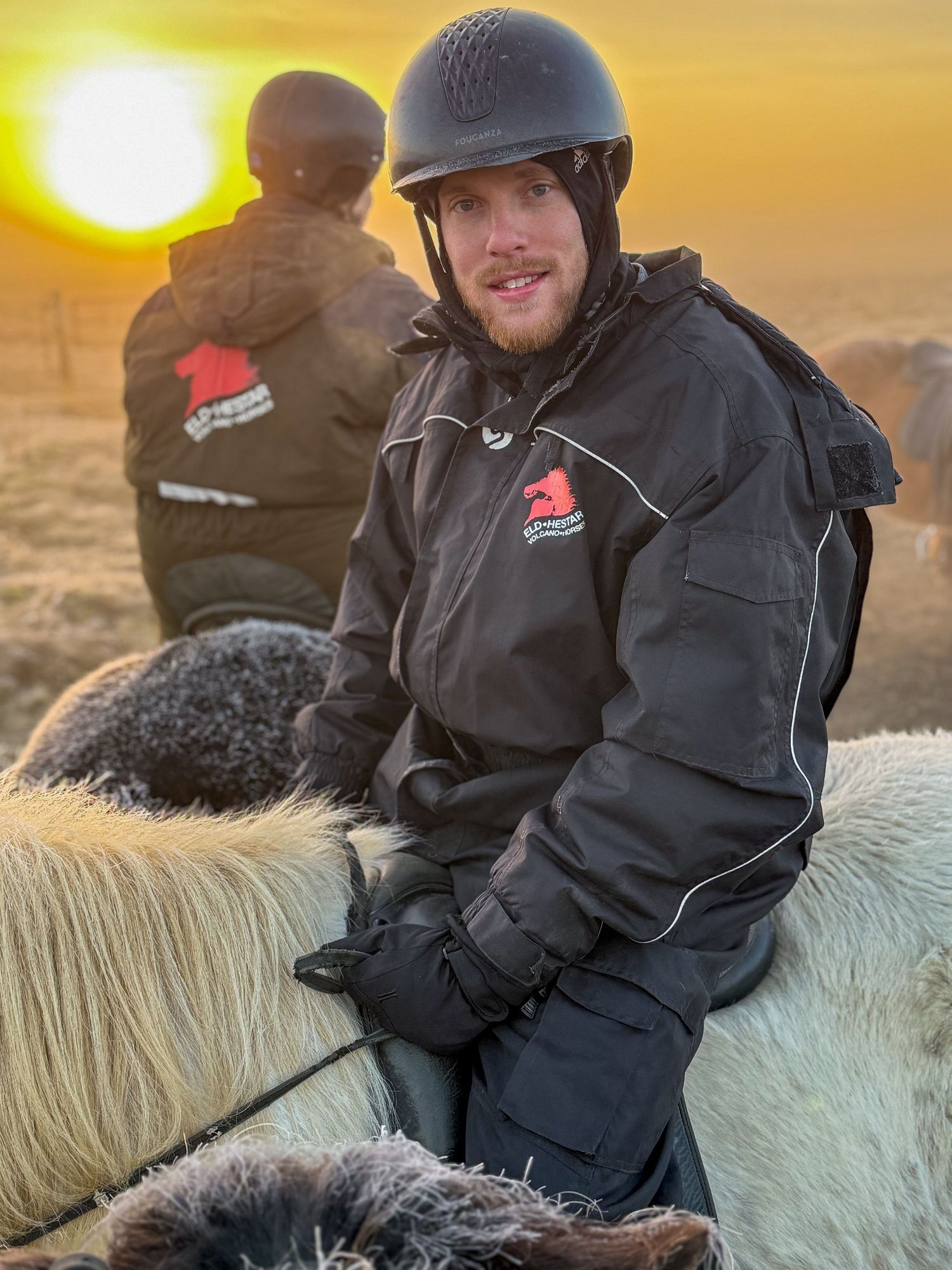 Person in dark winter clothing rides a white horse against a dramatic golden sunset sky in the background.