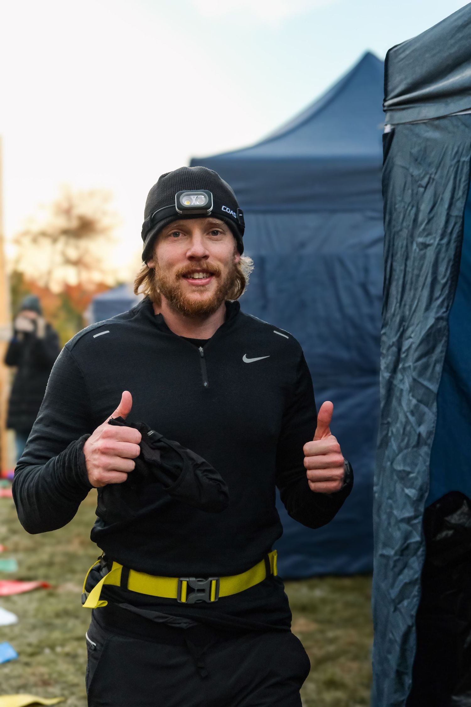 Person in black athletic wear and beanie gives enthusiastic thumbs up gestures outdoors near blue tent structures.
