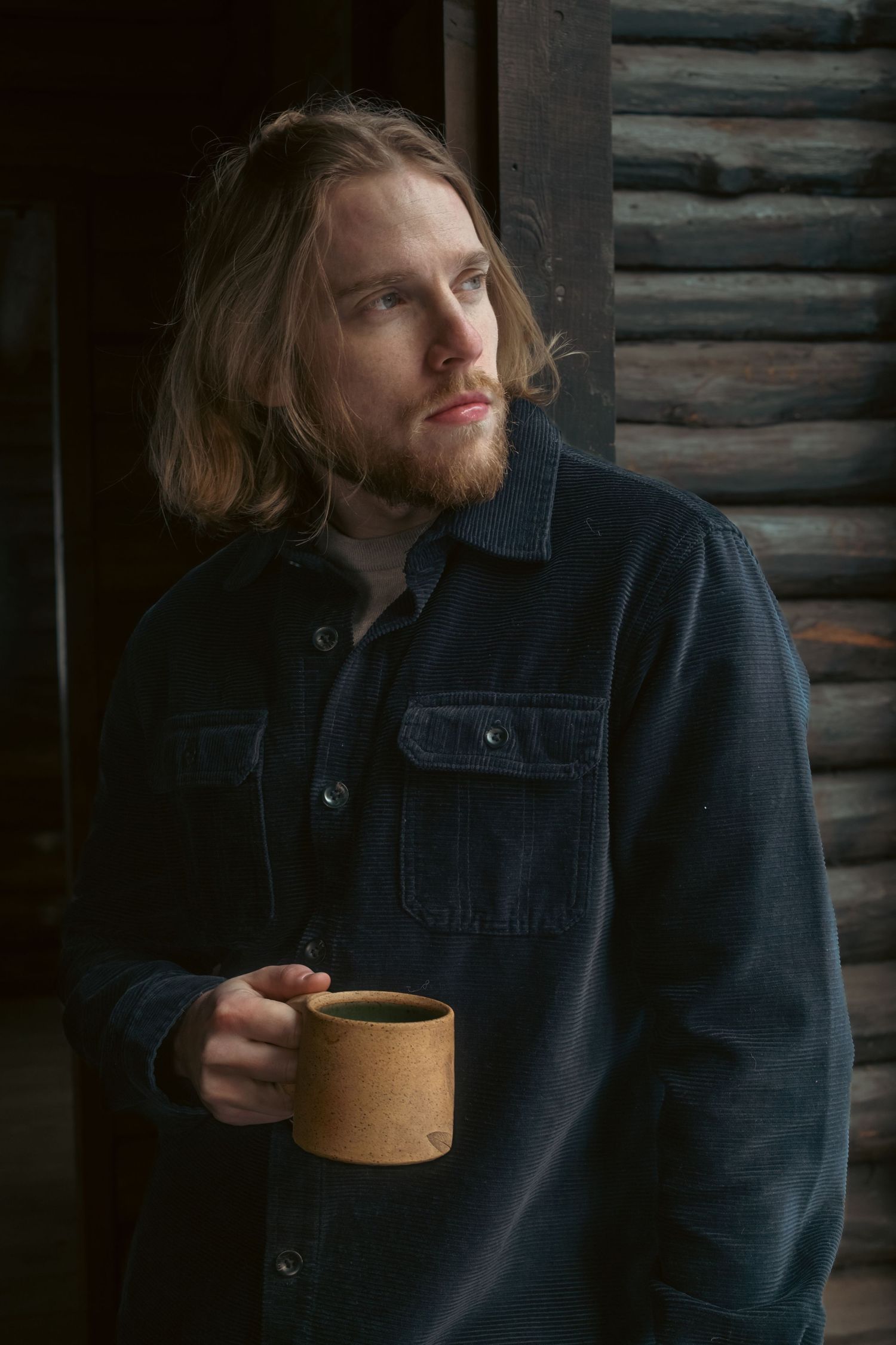 A person in a dark navy jacket sits in front of rustic wooden walls while drinking from a tan ceramic mug.