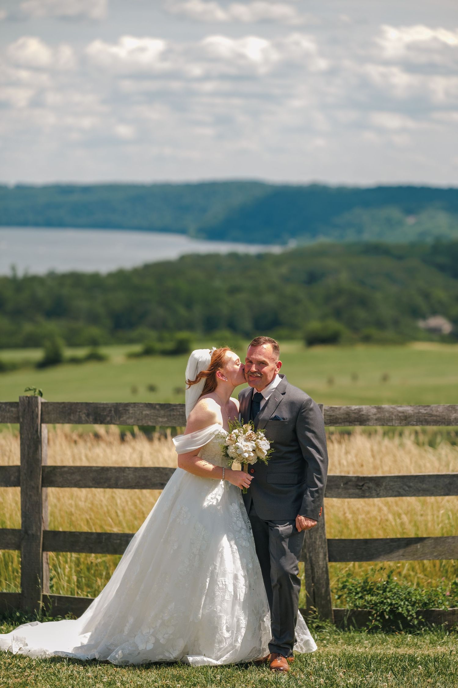 Bride and groom pose by rustic fence with scenic countryside lake view on sunny wedding day.
