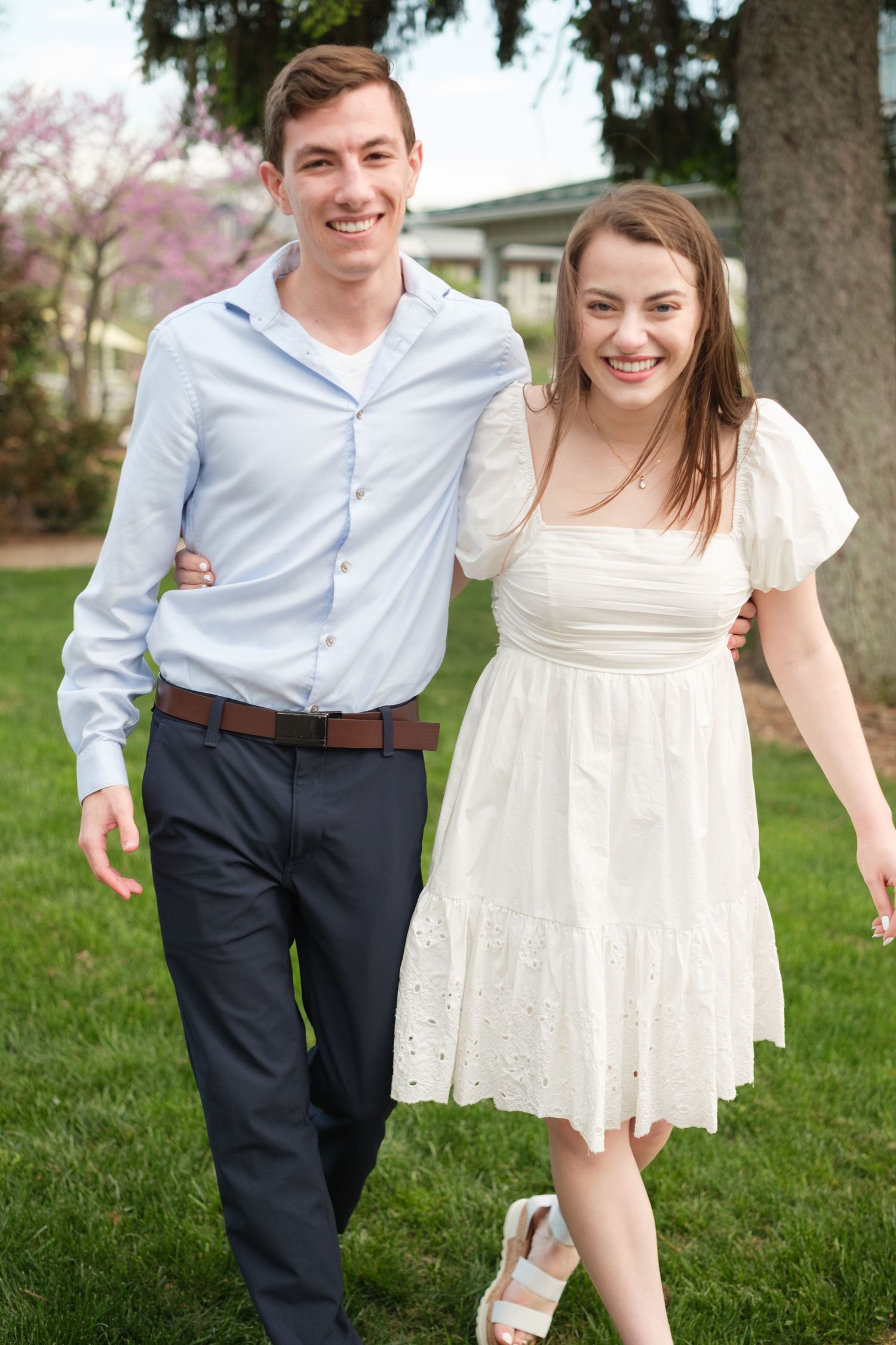 Young couple smiling and walking together outdoors on green grass, man in blue shirt and woman in white dress.