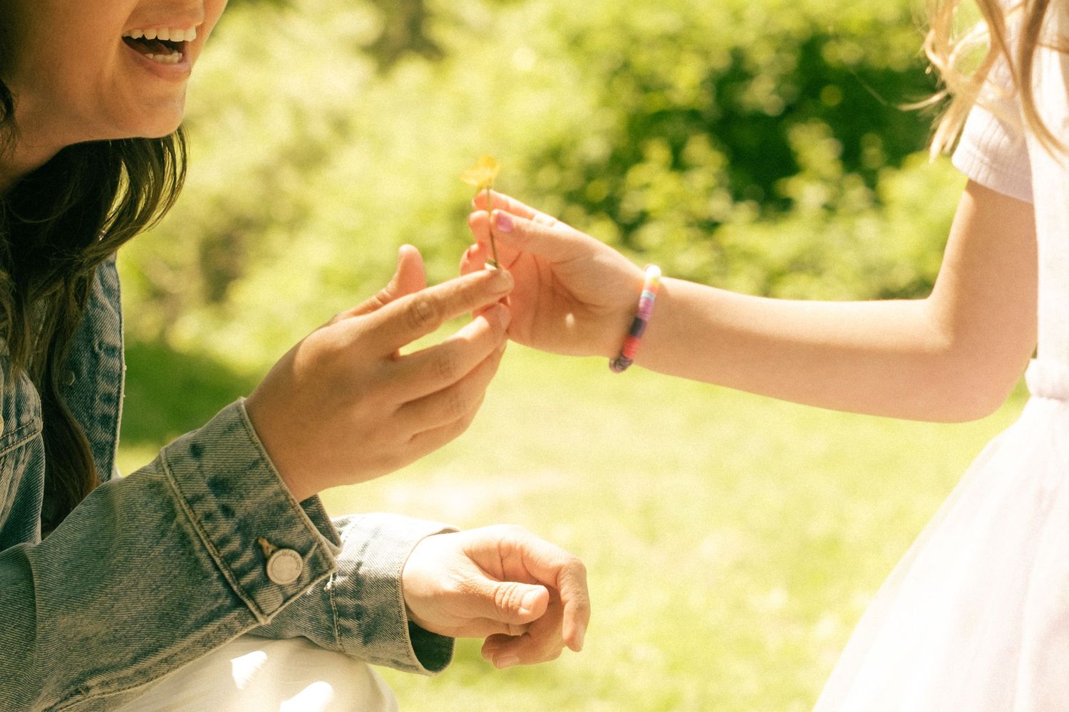 A woman in a denim jacket receives a small flower from a child in a white dress outdoors in a sunny green park.