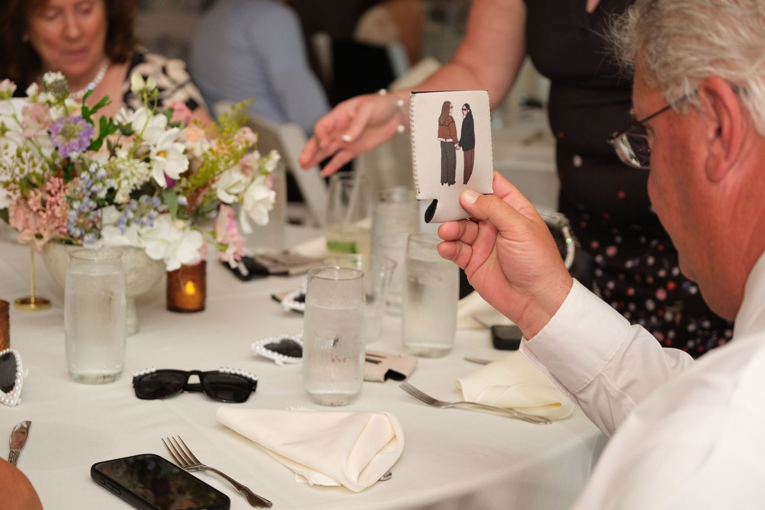 Guest holds a wedding photo card at a floral-decorated reception table with glassware and sunglasses nearby.