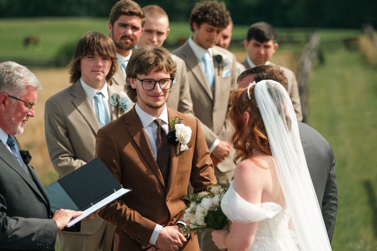 Groom in brown suit exchanges vows with red-haired bride at outdoor wedding ceremony with groomsmen.
