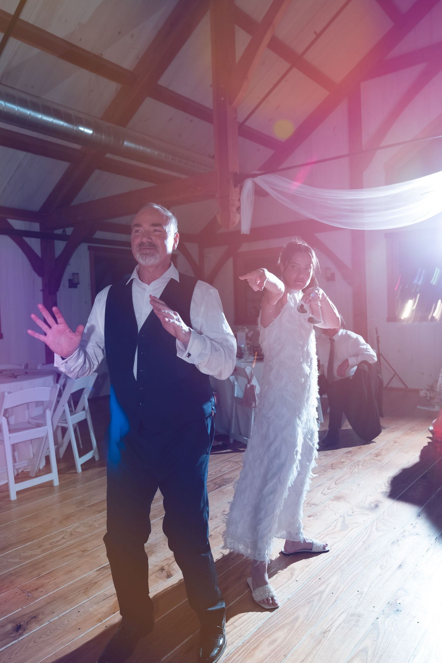 Older man in vest dancing joyfully at wedding reception in rustic barn venue with guests behind him.