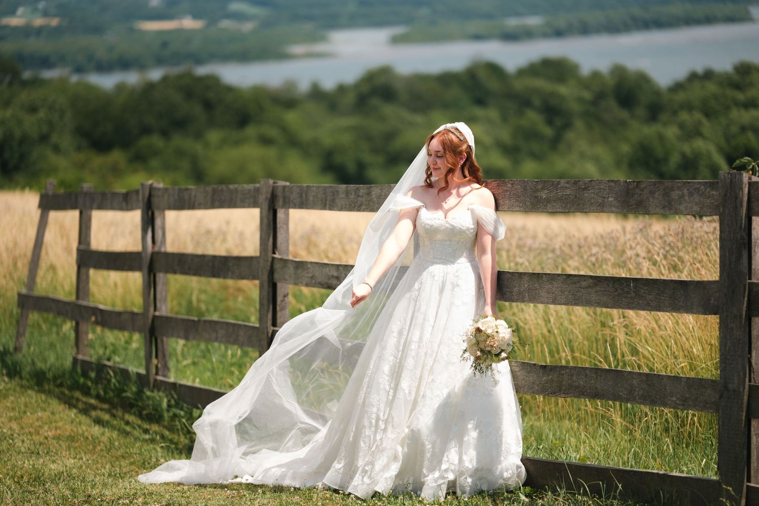 Bride in white lace gown and veil holds bouquet beside rustic wooden fence in sunny green meadow.