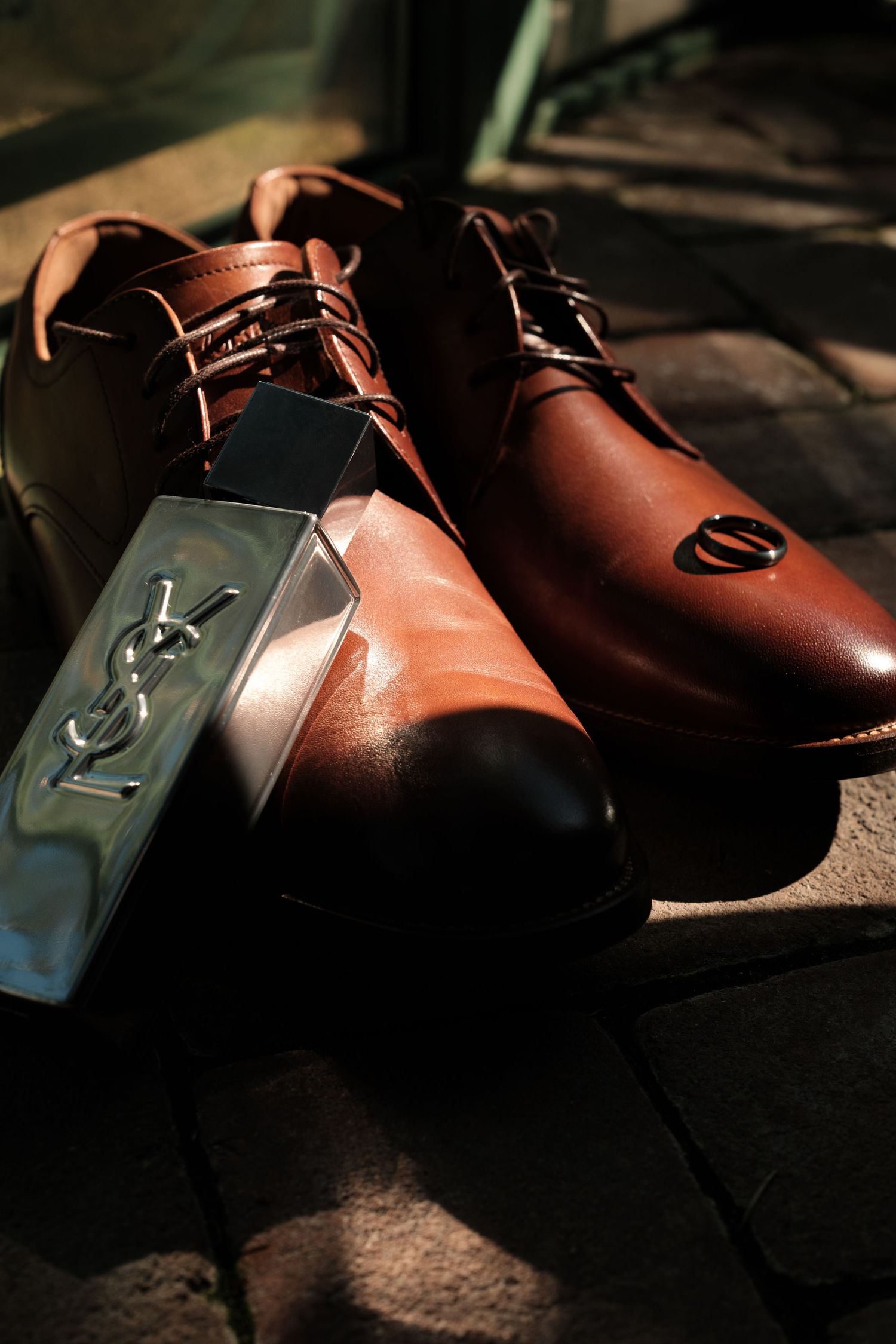 Pair of polished brown leather lace-up dress shoes on a brick surface with a YSL cologne bottle and a ring resting on one toe.