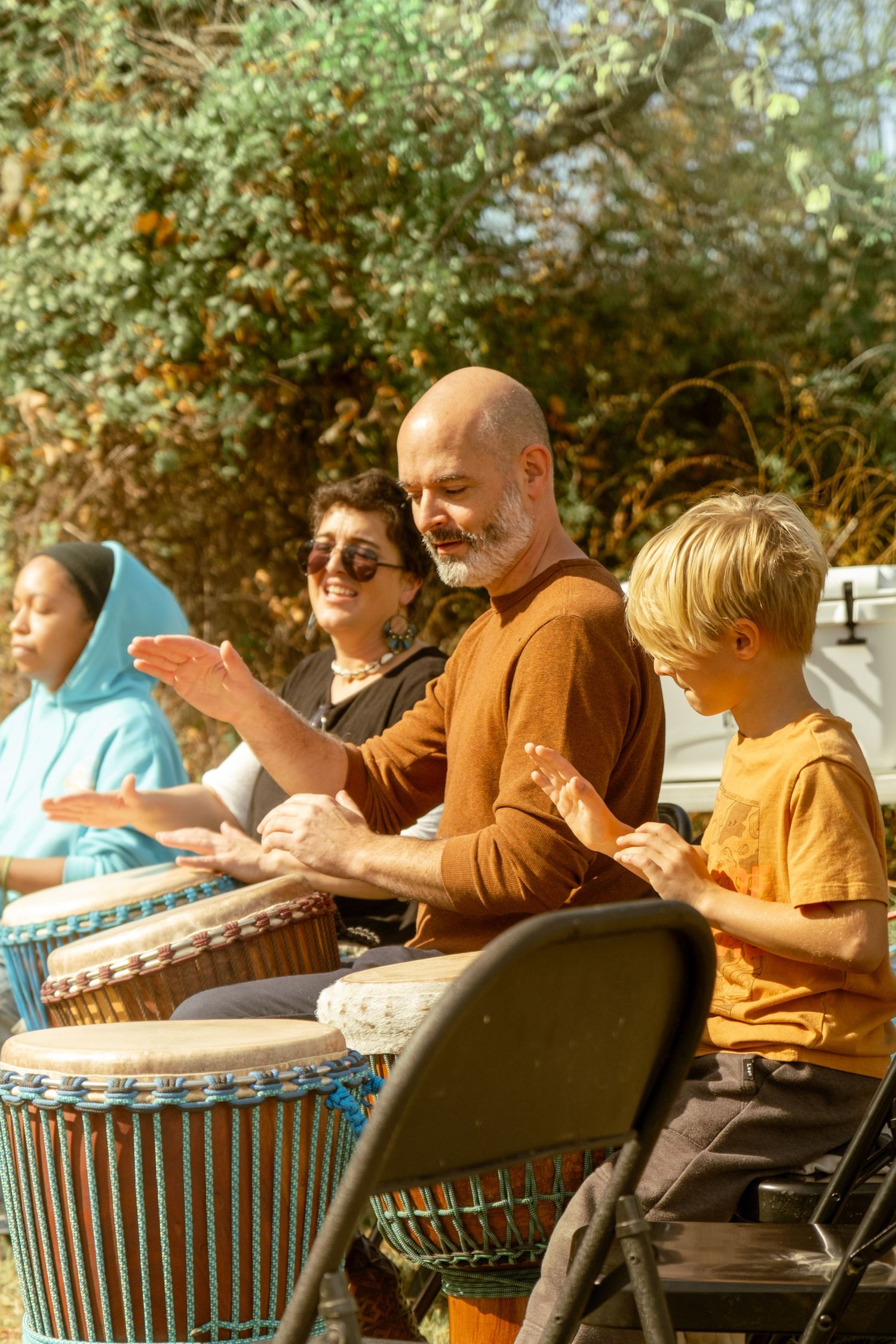 A group of people sit outdoors playing African djembe drums together in a drumming circle on a sunny day.