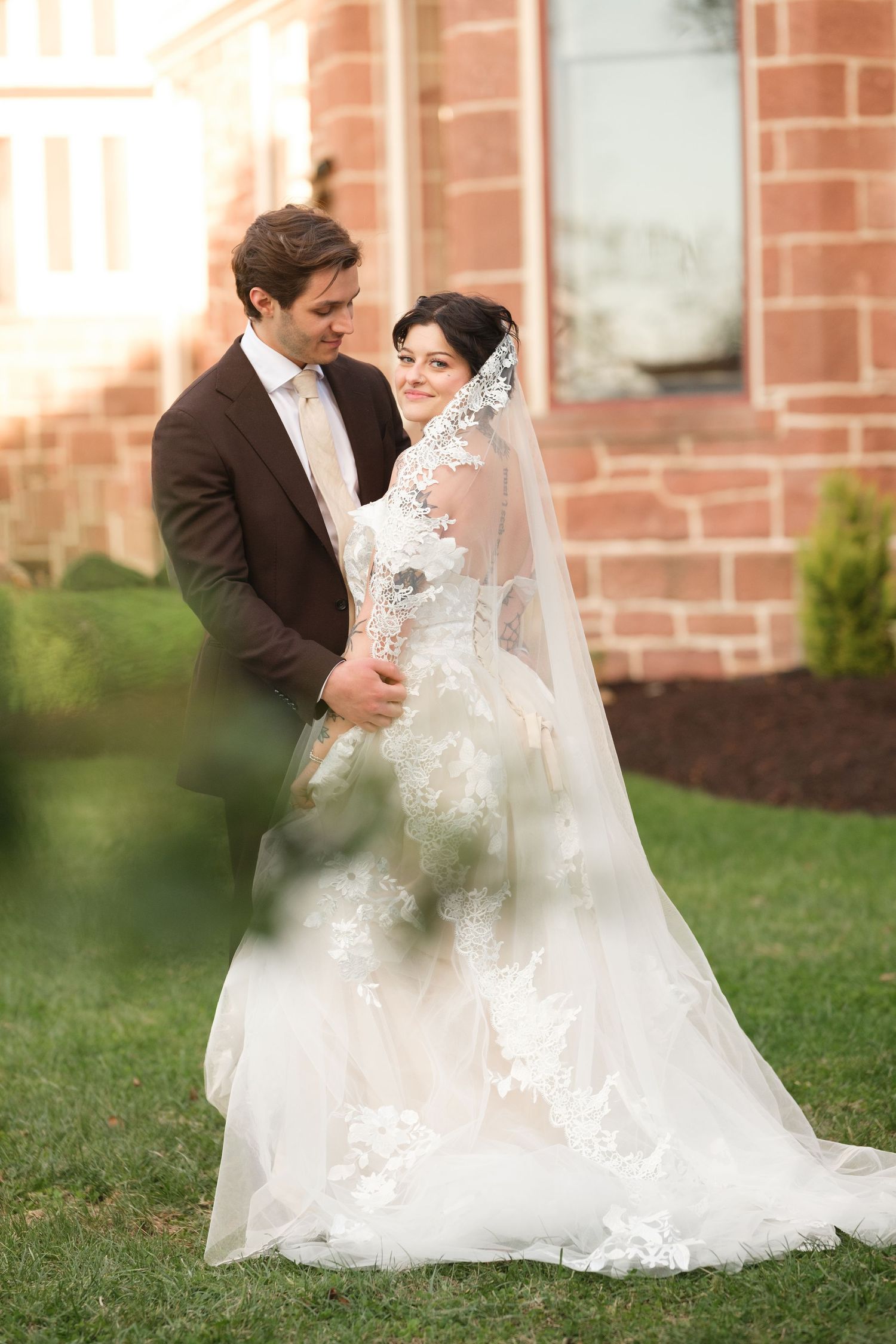 Bride in lace wedding gown and veil stands with groom in brown suit on green lawn beside brick building.