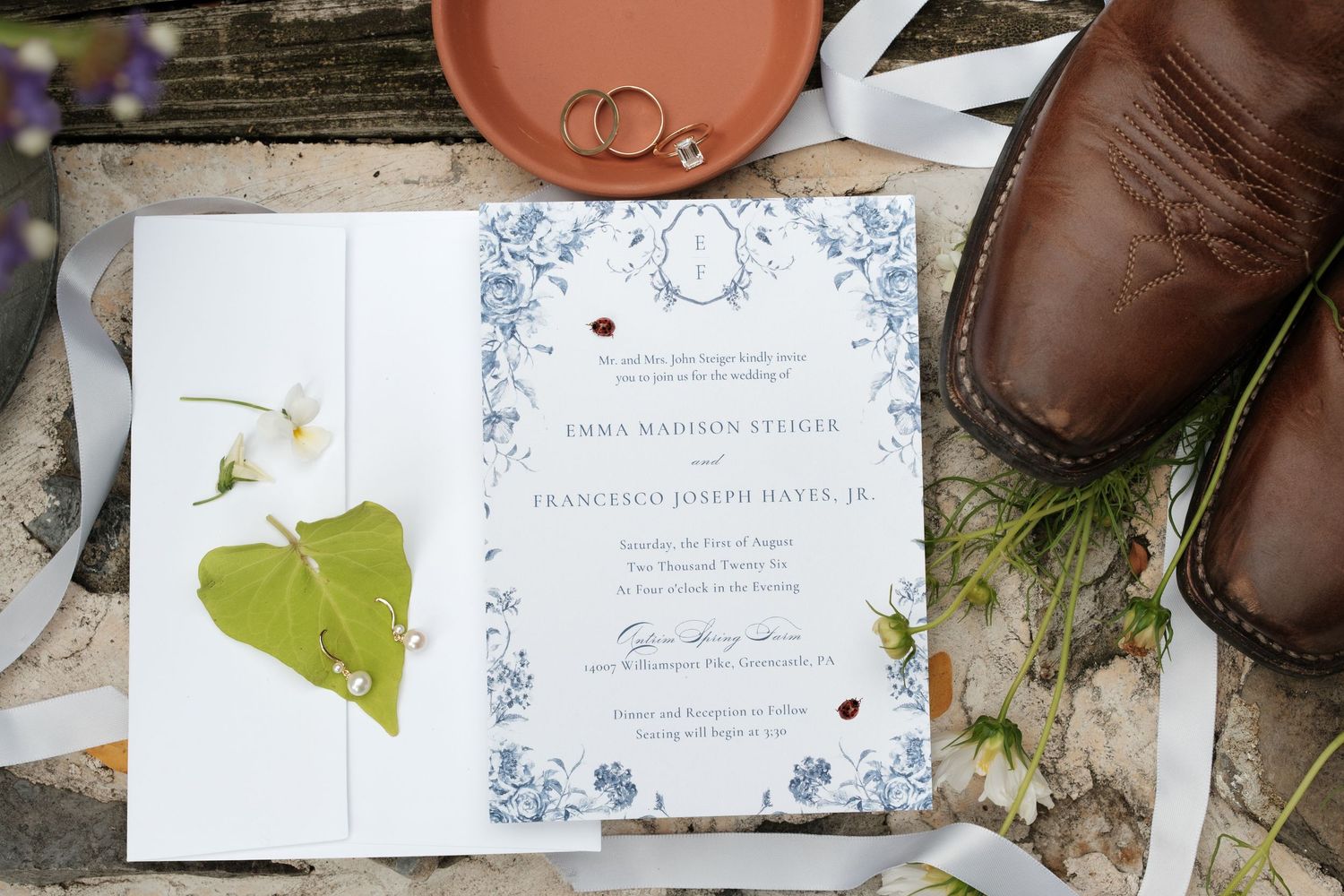 Elegant wedding invitation with blue floral border, surrounded by rings, brown leather shoes, ribbon, and wildflowers on rustic wood.