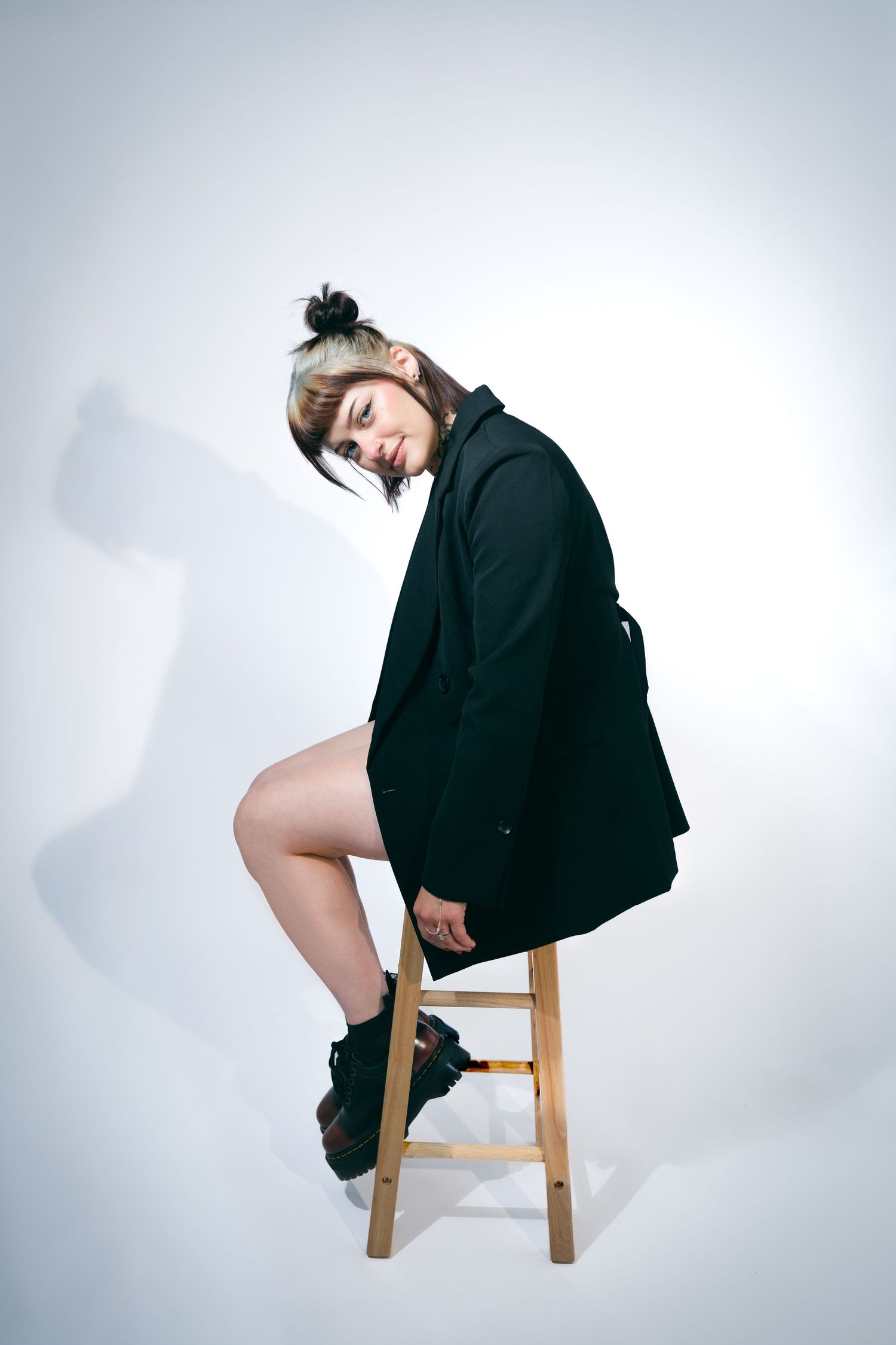 Woman with a top knot and bangs sits on a wooden stool in a black blazer and boots, tilting her head against a white studio backdrop.