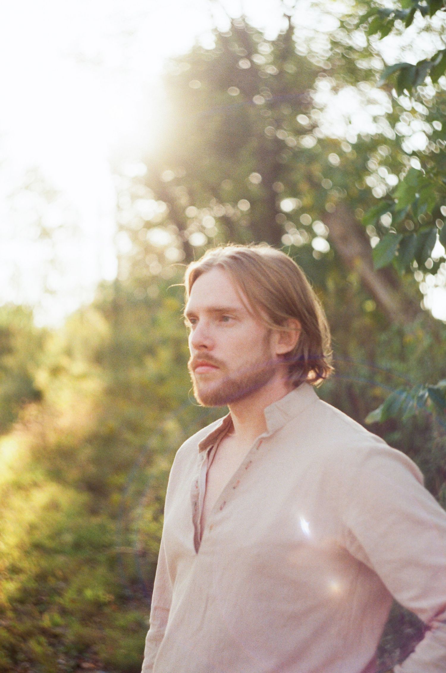 Portrait photo in soft sunlight showing person wearing white button-down shirt against blurred natural outdoor background.