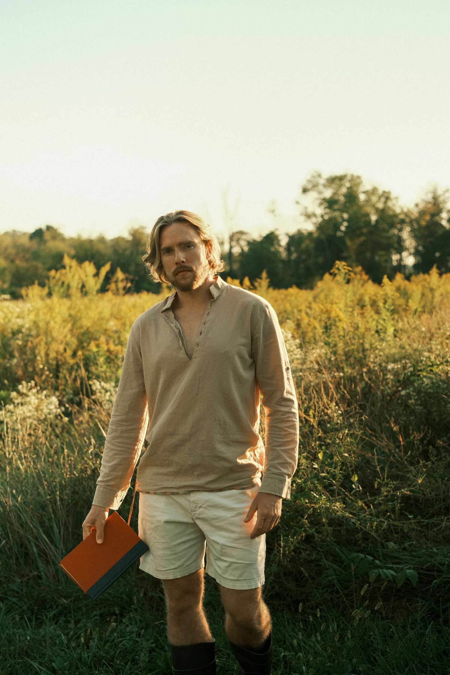 Man holding orange book standing in sunlit field with wildflowers and trees in background during golden hour.