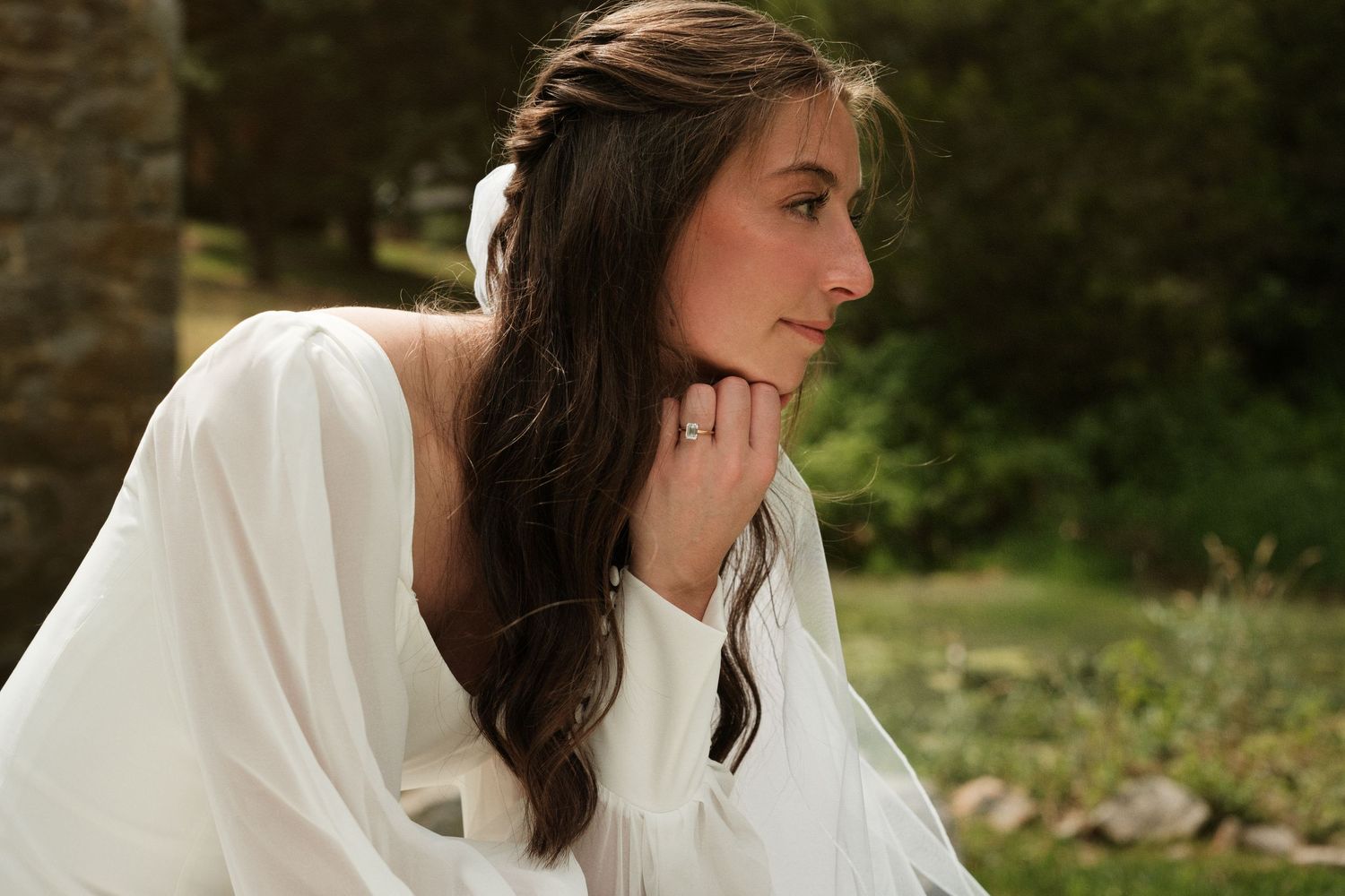 Young woman in white dress sitting outdoors, gazing thoughtfully with hand near chin, surrounded by lush greenery.