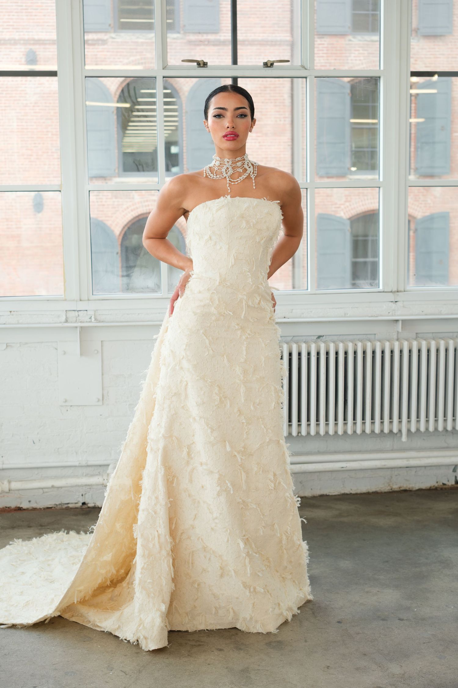 Model wearing a strapless ivory A-line wedding gown with floral texture, a statement necklace, posing in a bright studio.