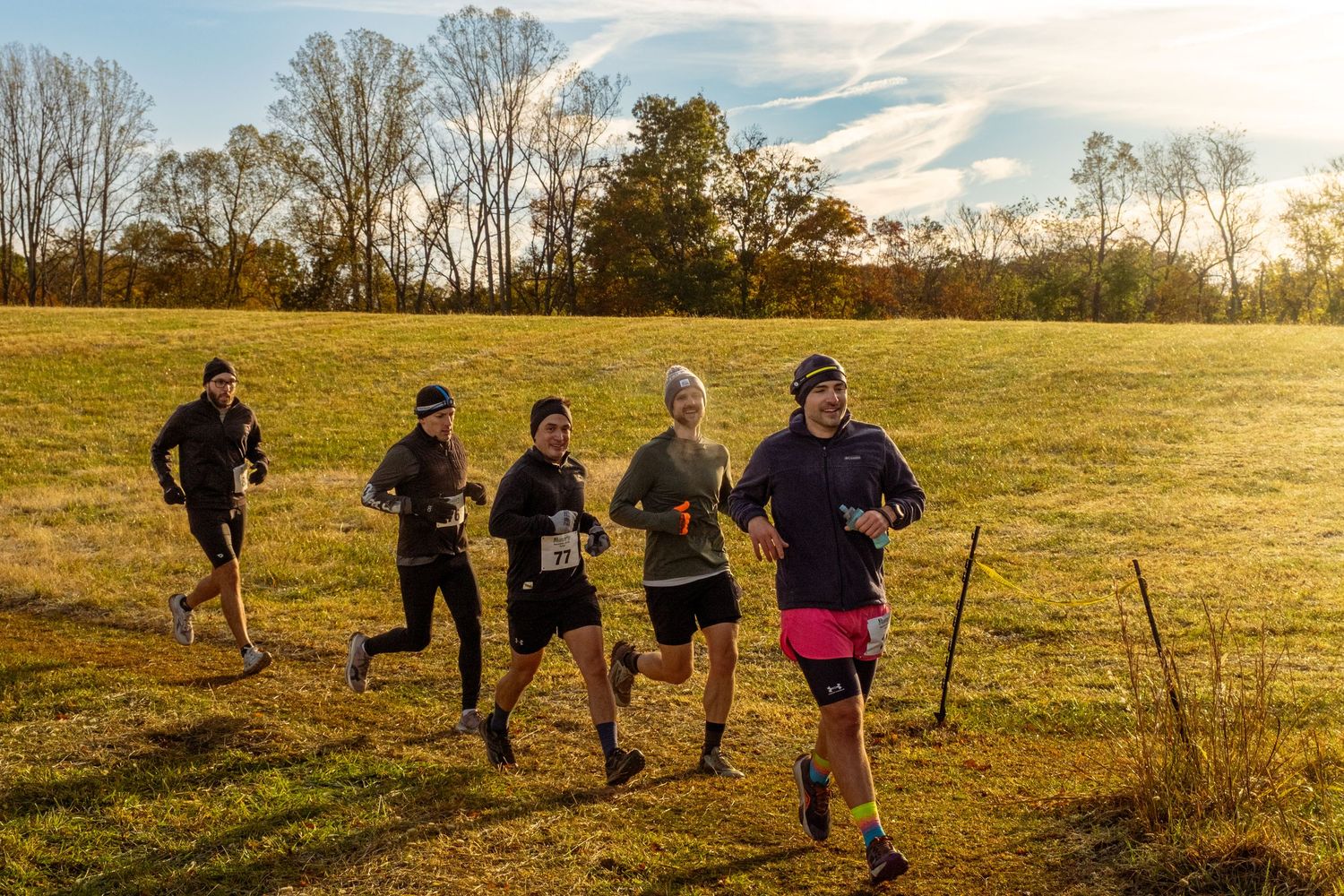 Cross country runners traverse an open field during golden hour as the sun sets behind bare trees in autumn.