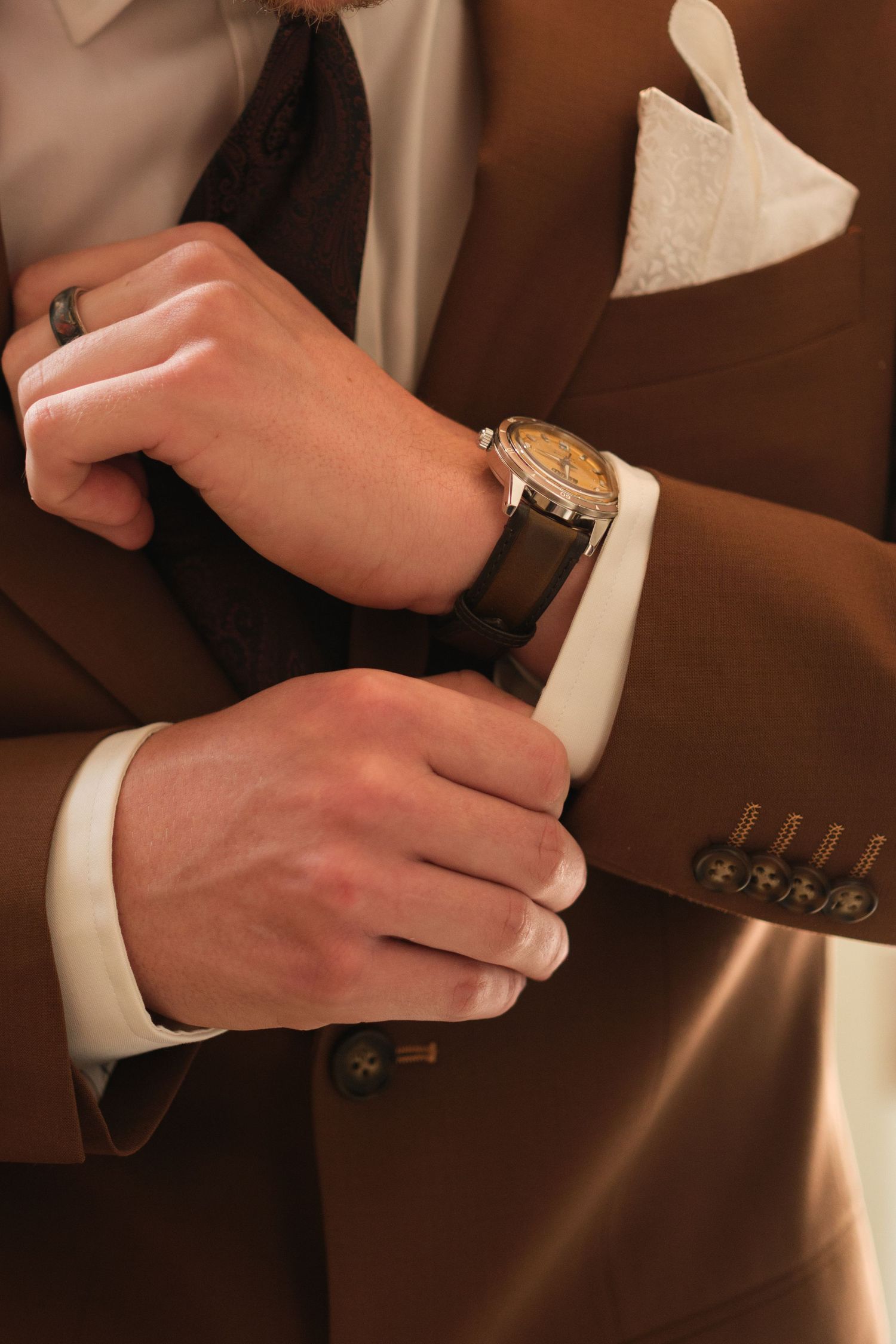 Man in brown suit adjusting cufflinks, wearing a classic wristwatch and white pocket square.