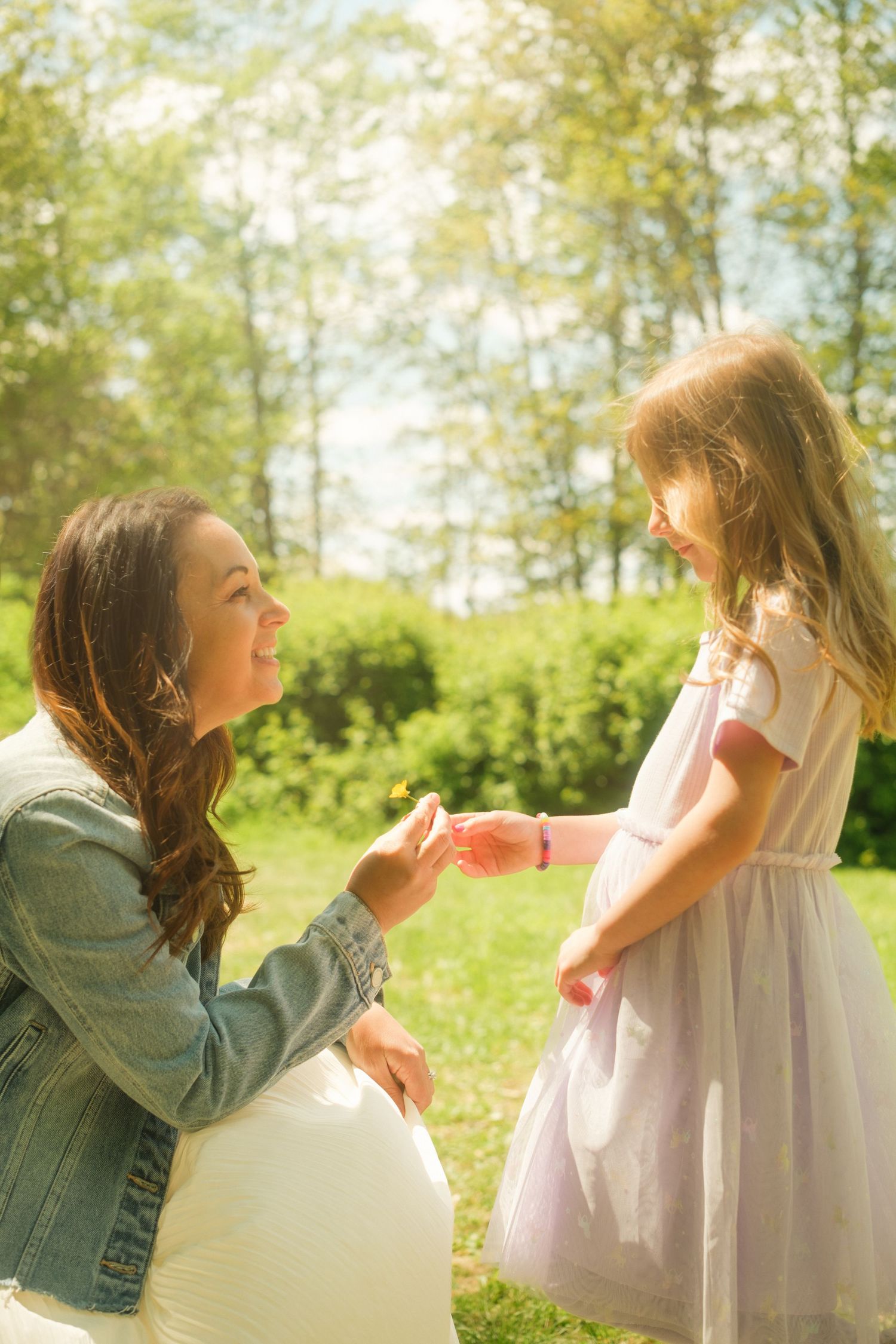 Pregnant woman in denim jacket kneeling outdoors, smiling at young girl in pink dress in sunny green park.