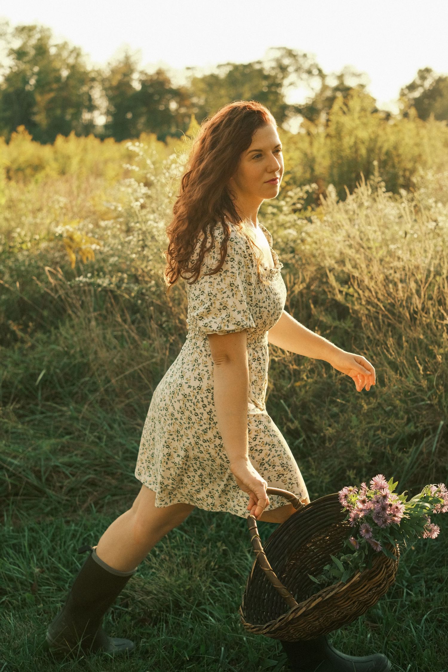 Woman in floral dress and green boots carrying a wicker basket with flowers through a sunlit meadow.