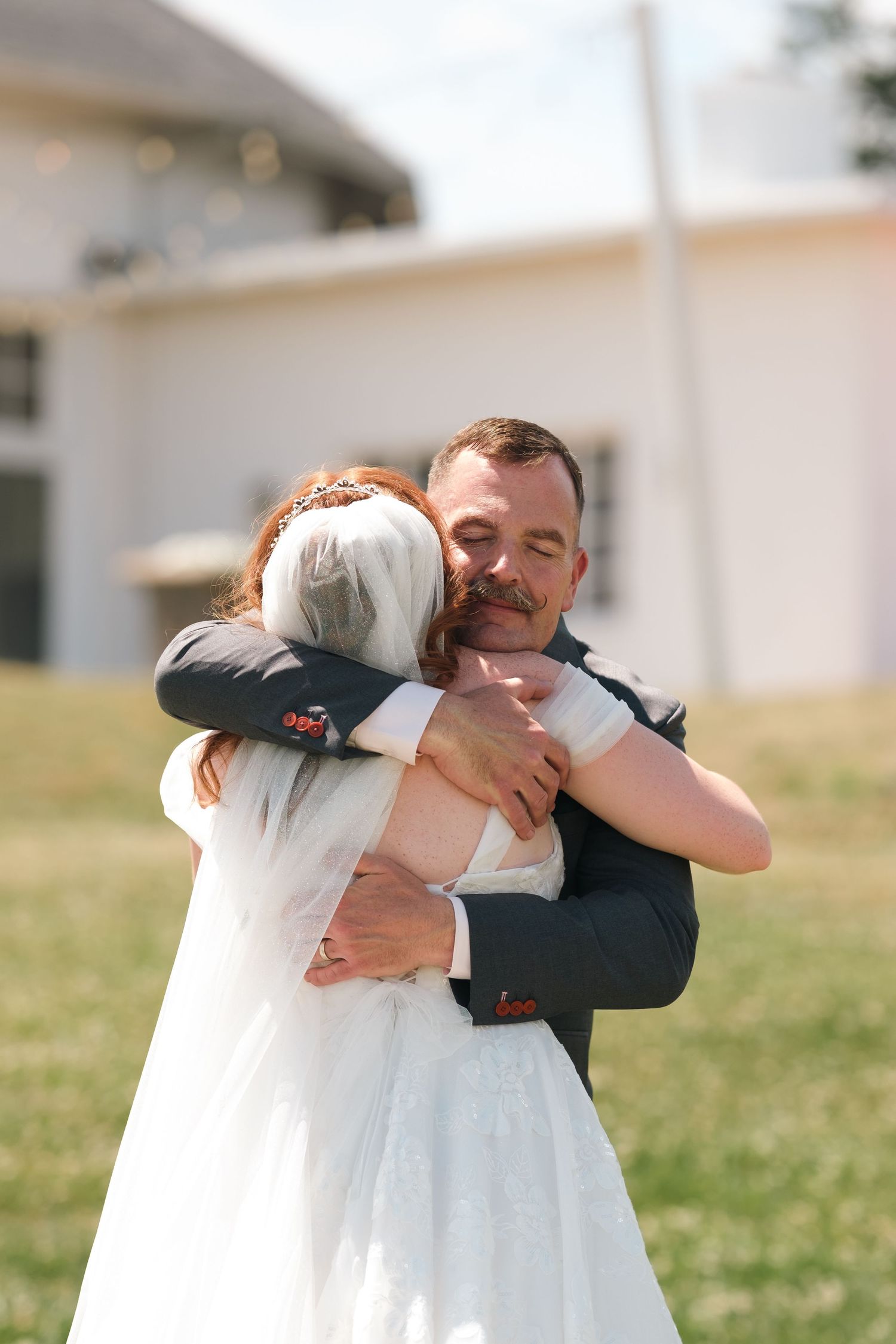 A groom in a dark suit embraces a bride in a white dress and veil outdoors on a sunny wedding day.