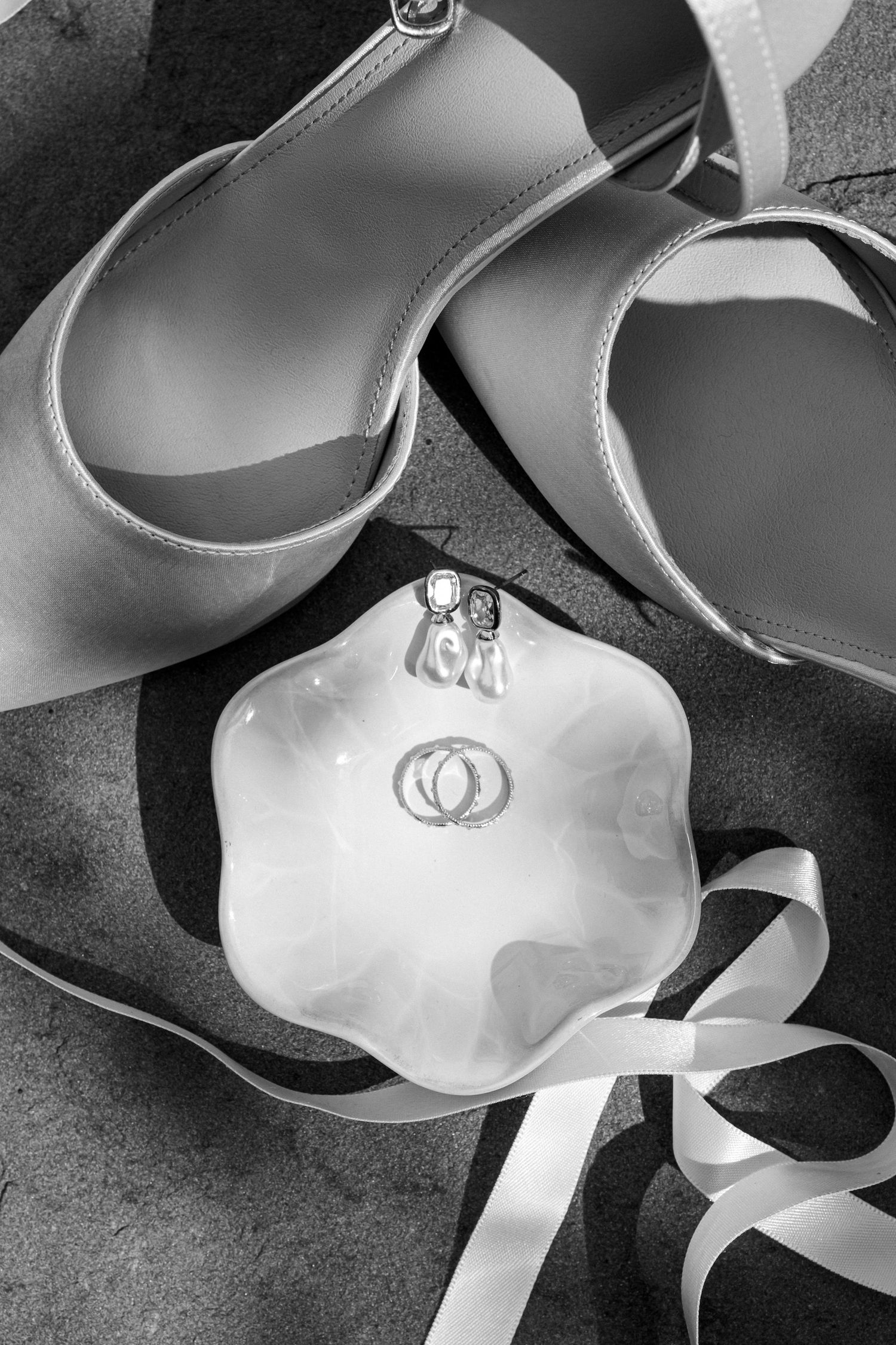 Black and white flat lay of wedding rings in a dish beside bridal heels and white ribbon.