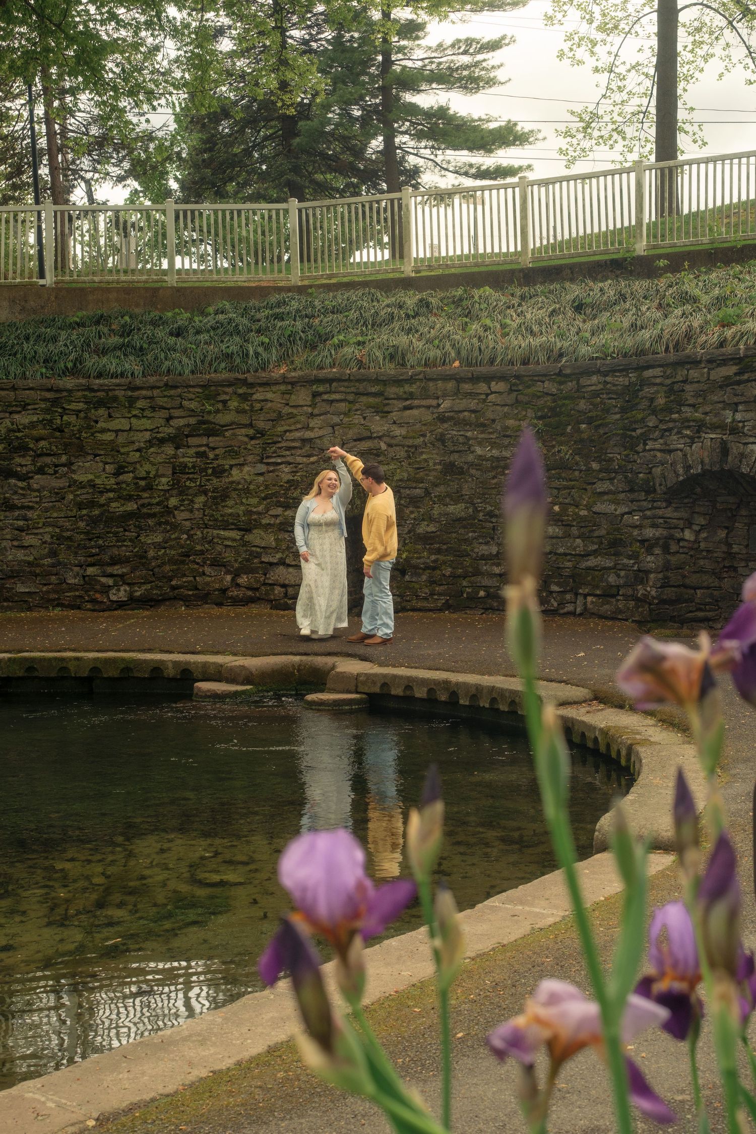 Couple dancing by a serene circular pond surrounded by stone walls, with purple iris flowers in the foreground.