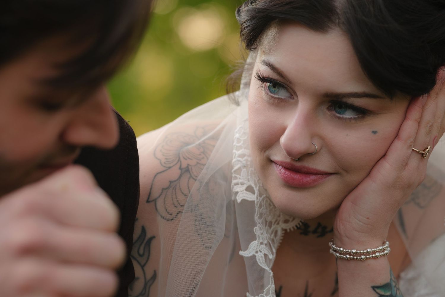 Tattooed bride in lace veil smiles softly at groom during intimate wedding portrait outdoors.
