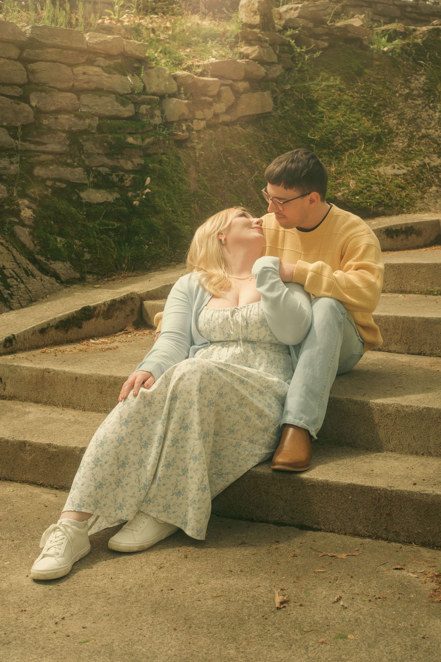 Engaged Couple sitting on stone steps, woman in floral dress leaning toward man in yellow sweater and jeans in romantic outdoor setting.