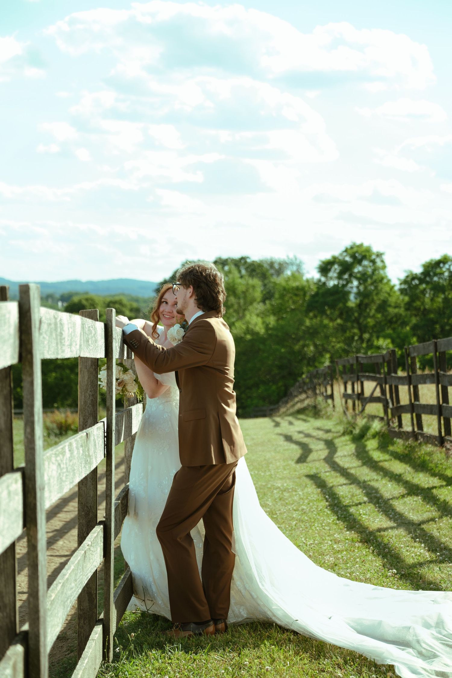 A bride and groom share a romantic kiss beside a rustic wooden fence on a sunny countryside farm.