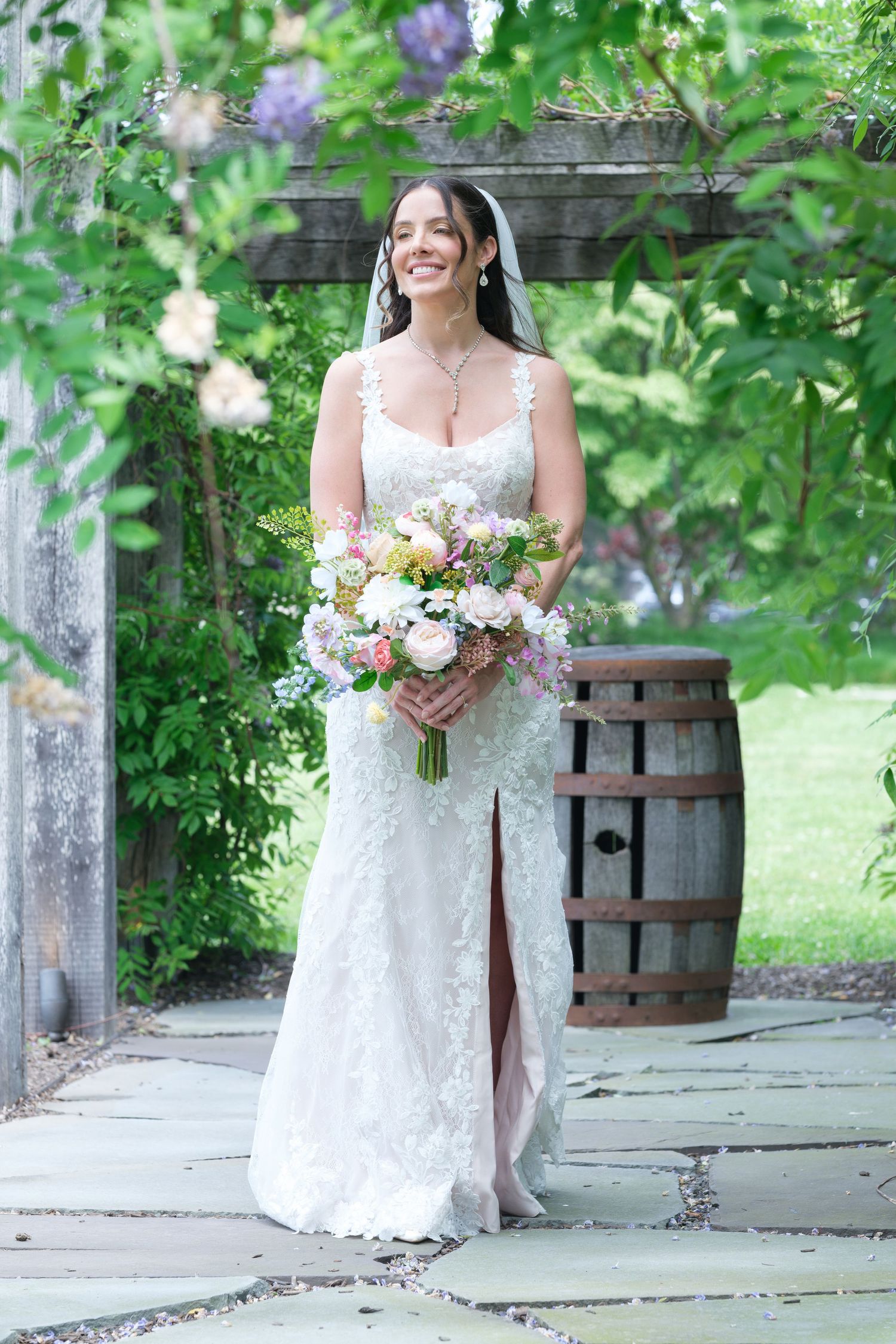 Smiling bride in lace wedding gown holding bouquet under floral garden arbor.